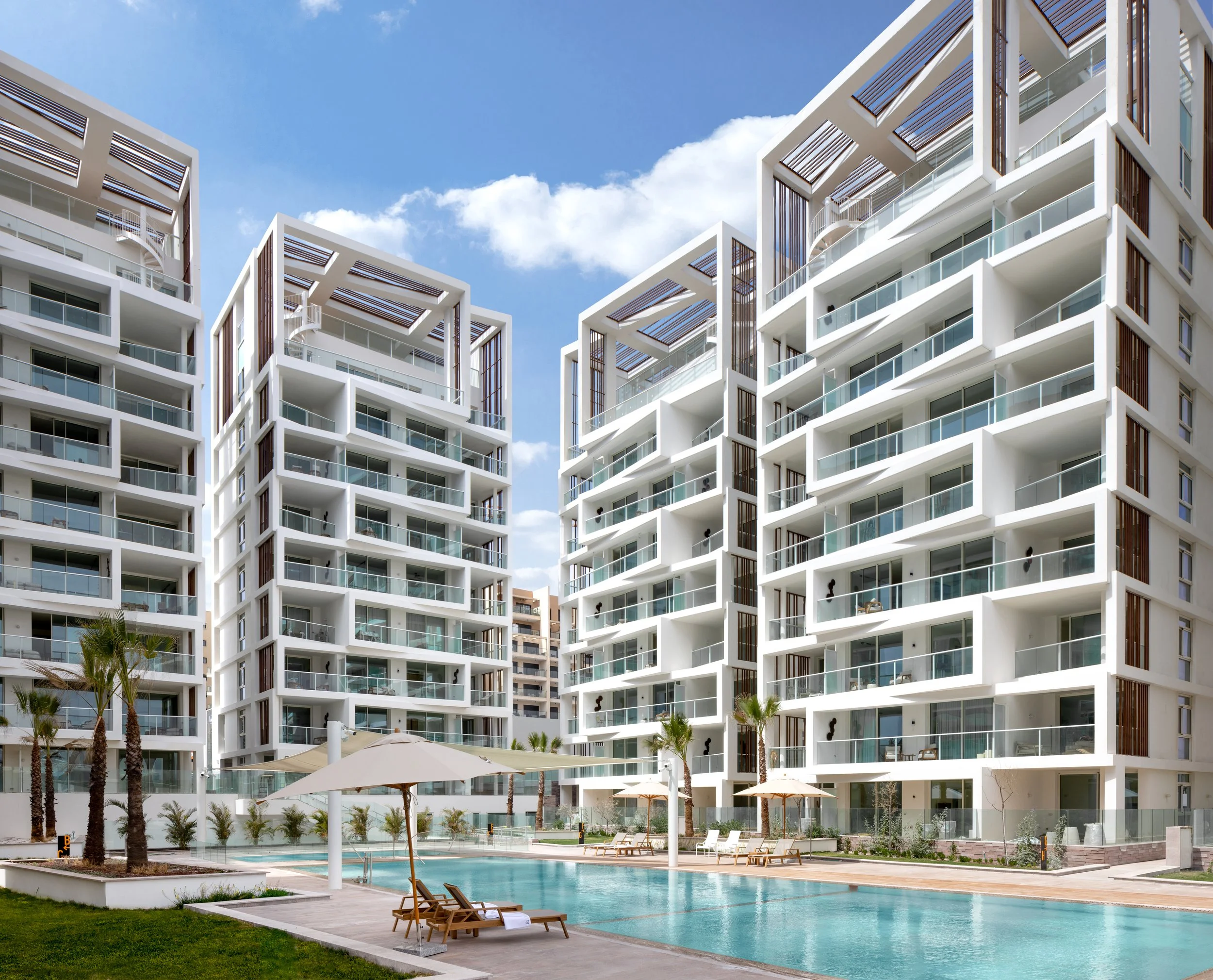 Modern white residential high-rise buildings with balconies surrounding a swimming pool area with lounge chairs and umbrellas, under a blue sky with clouds.
