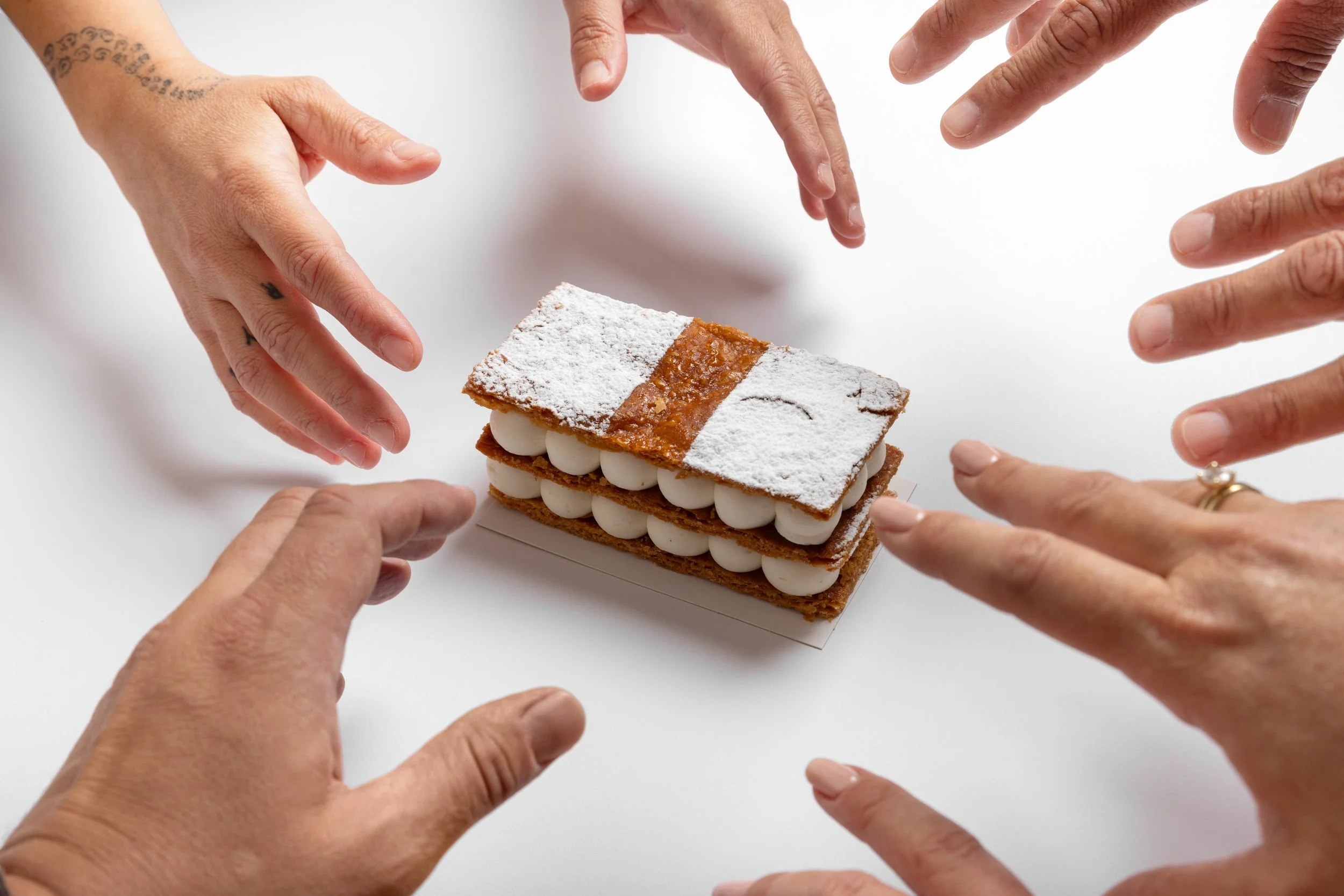 Multiple hands reaching towards a rectangular layered dessert with white filling, dusted with powdered sugar, on a white background.