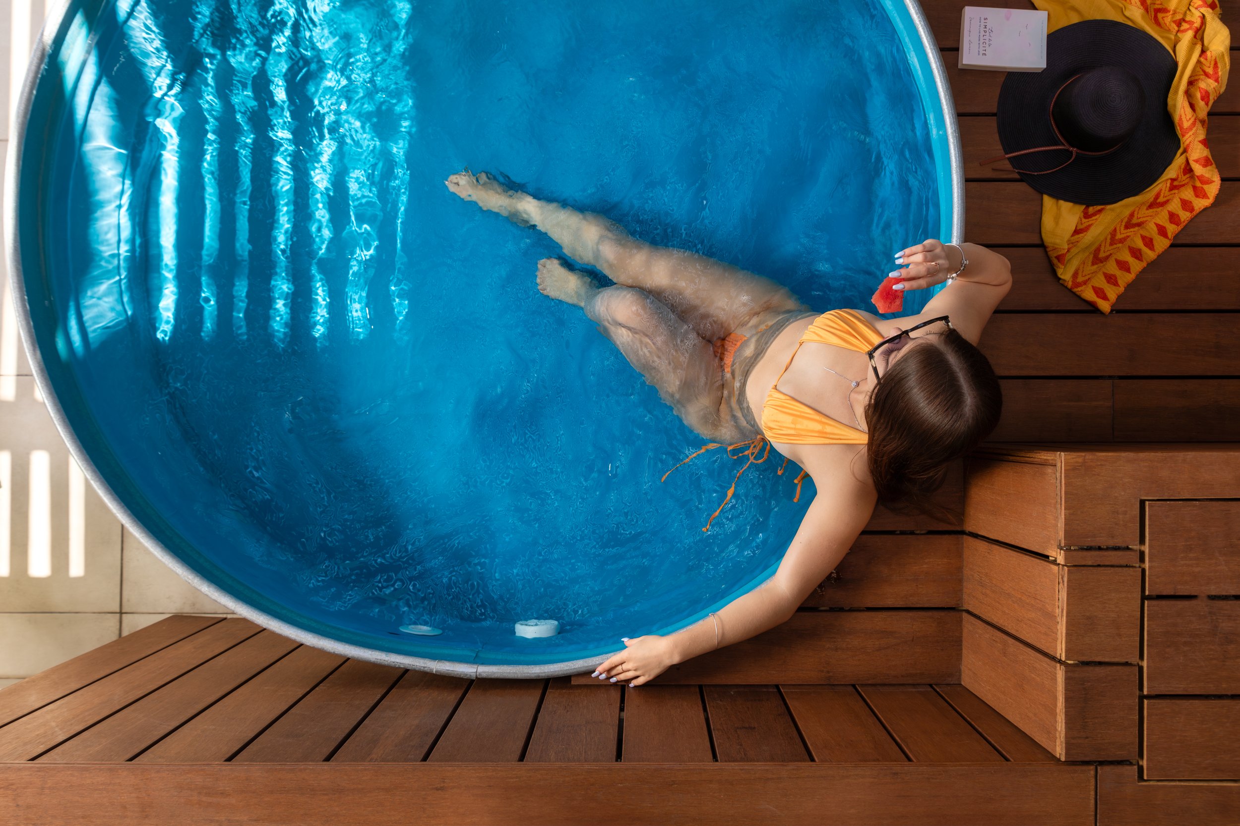 Woman in an orange swimsuit lounging in a small round pool, holding a slice of watermelon, on a wooden deck with hat and towel nearby.
