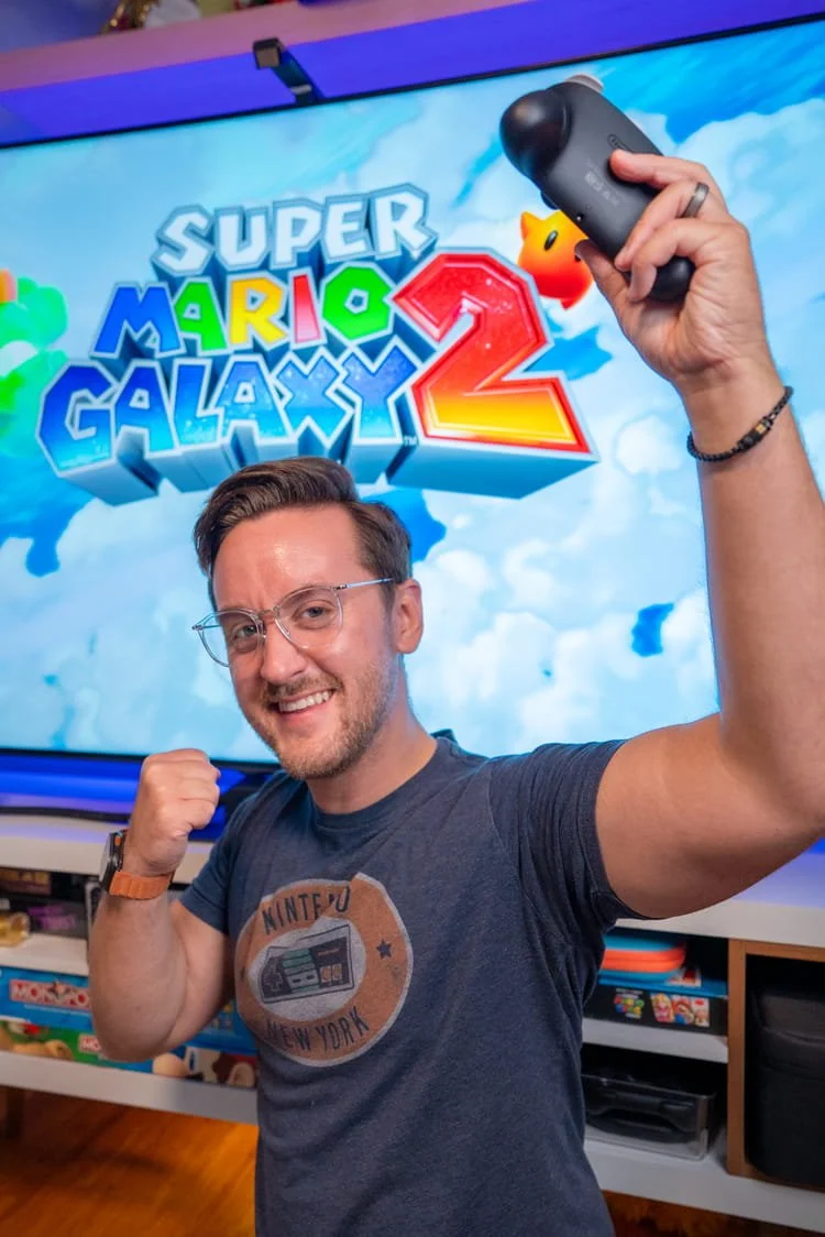 A man smiling and flexing his arm in front of a TV screen displaying the Super Mario Galaxy 2 video game logo, holding a gaming controller in his raised hand.