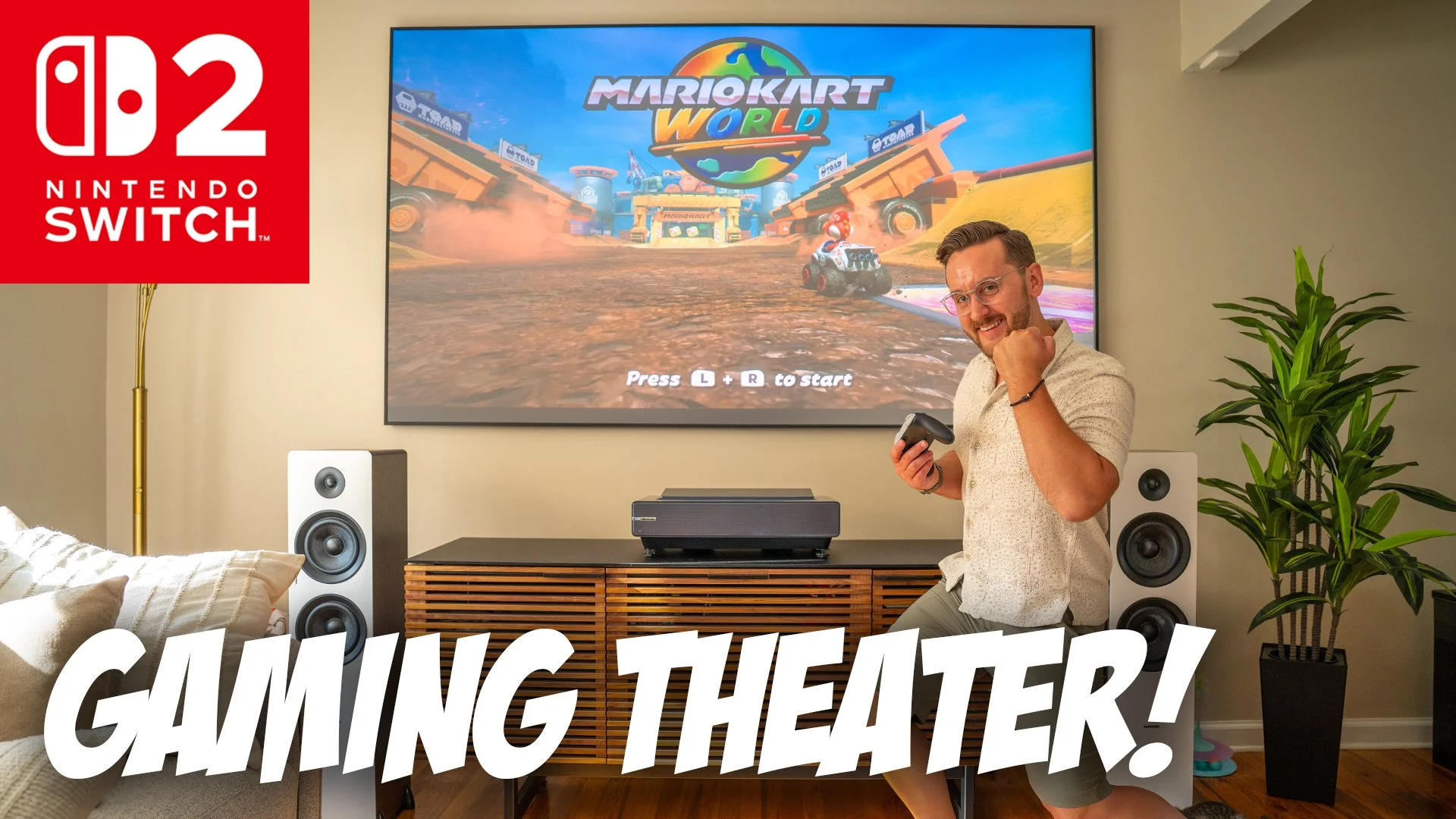 A man sitting in a living room gaming in front of a large TV screen displaying the game Mario Kart World on Nintendo Switch. The room has a modern entertainment setup with speakers, a console, and a decorative cabinet. There is a gross entertainment poster logo in the top left corner and bold text at the bottom reading 'GAMING THEATER!'