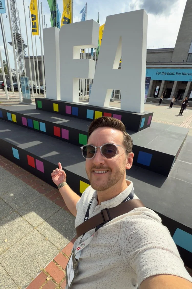 A man wearing sunglasses and a beige shirt taking a selfie in front of large white letters 'FA' at an outdoor event.