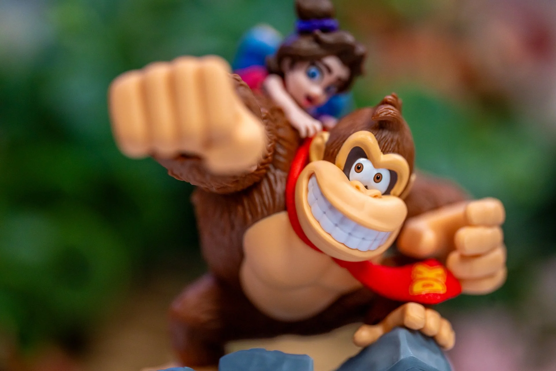Close-up of Donkey Kong toy figure with a wide grin, wearing a red tie, with Mabel Pines figure from Gravity Falls riding on his back, set against a blurred green and red background.