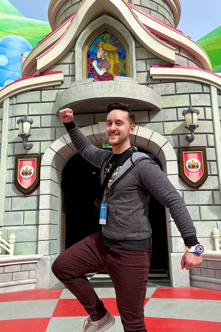 A man smiling and posing confidently with a raised fist in front of a castle themed structure at an amusement park, featuring cartoon shields and colorful murals.