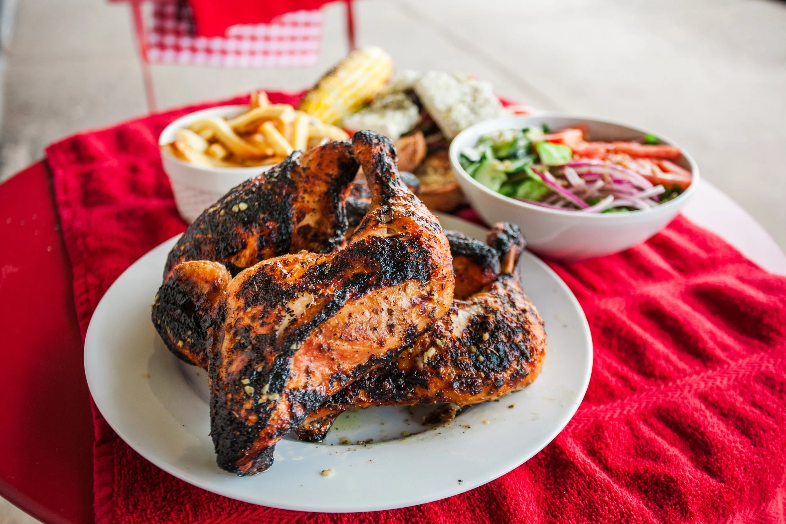 Grilled whole chicken on a white plate with side dishes including coleslaw, French fries, and grilled vegetables on a red tablecloth.