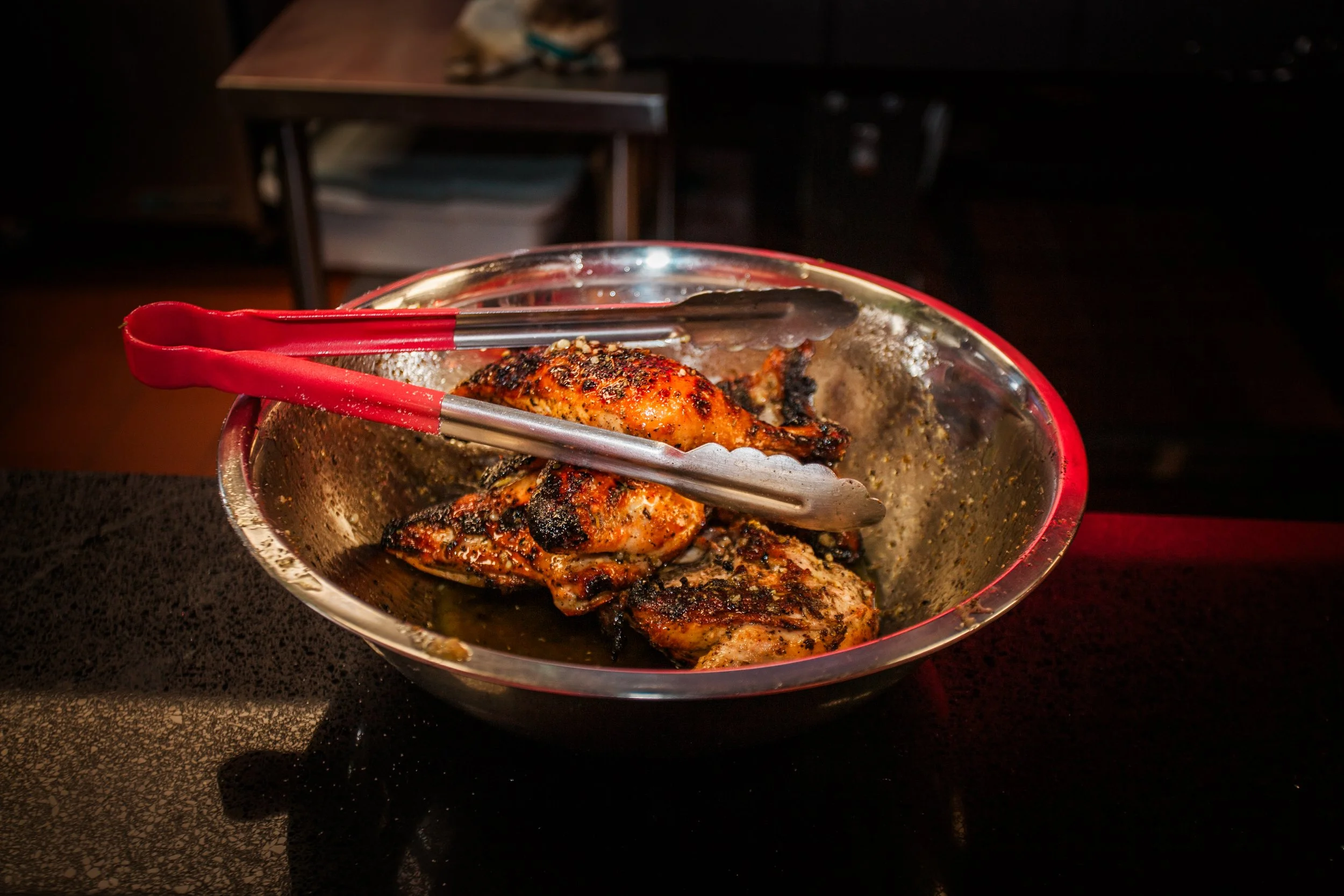 Grilled chicken pieces in a metal bowl with red tongs resting on top, set on a dark countertop.