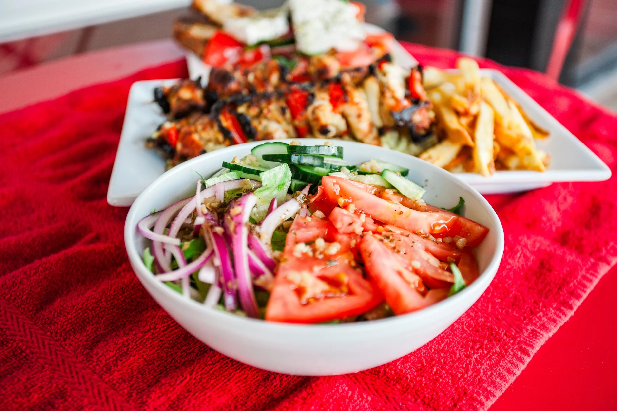 Fresh salad with sliced tomatoes, cucumbers, red onions, and lettuce in a white bowl, served on a red tablecloth. In the background, there are plates of grilled chicken and pasta.
