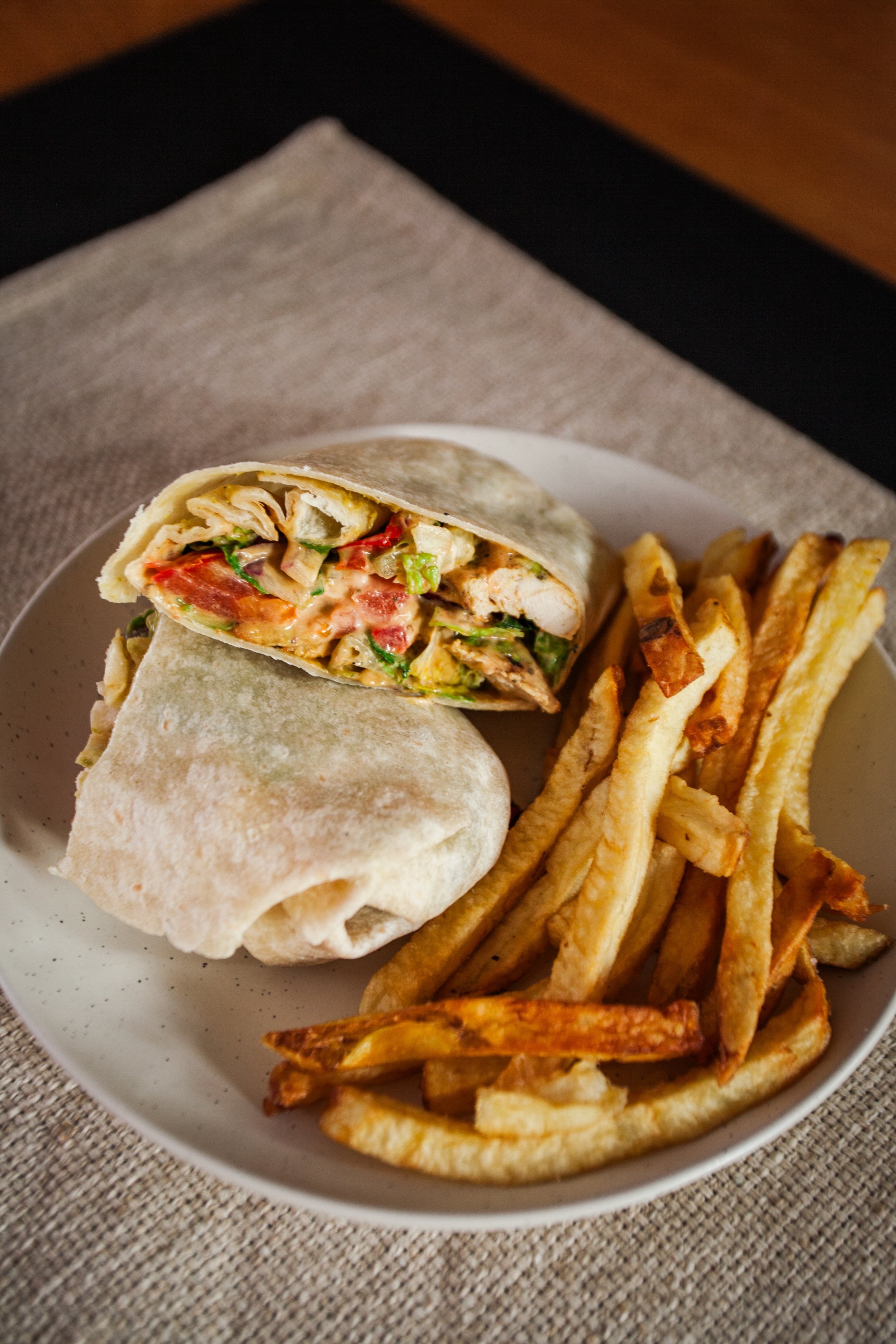 A plate with a chicken wrap cut in half, showing chicken, lettuce, tomatoes, and sauce, served with crinkle-cut French fries.
