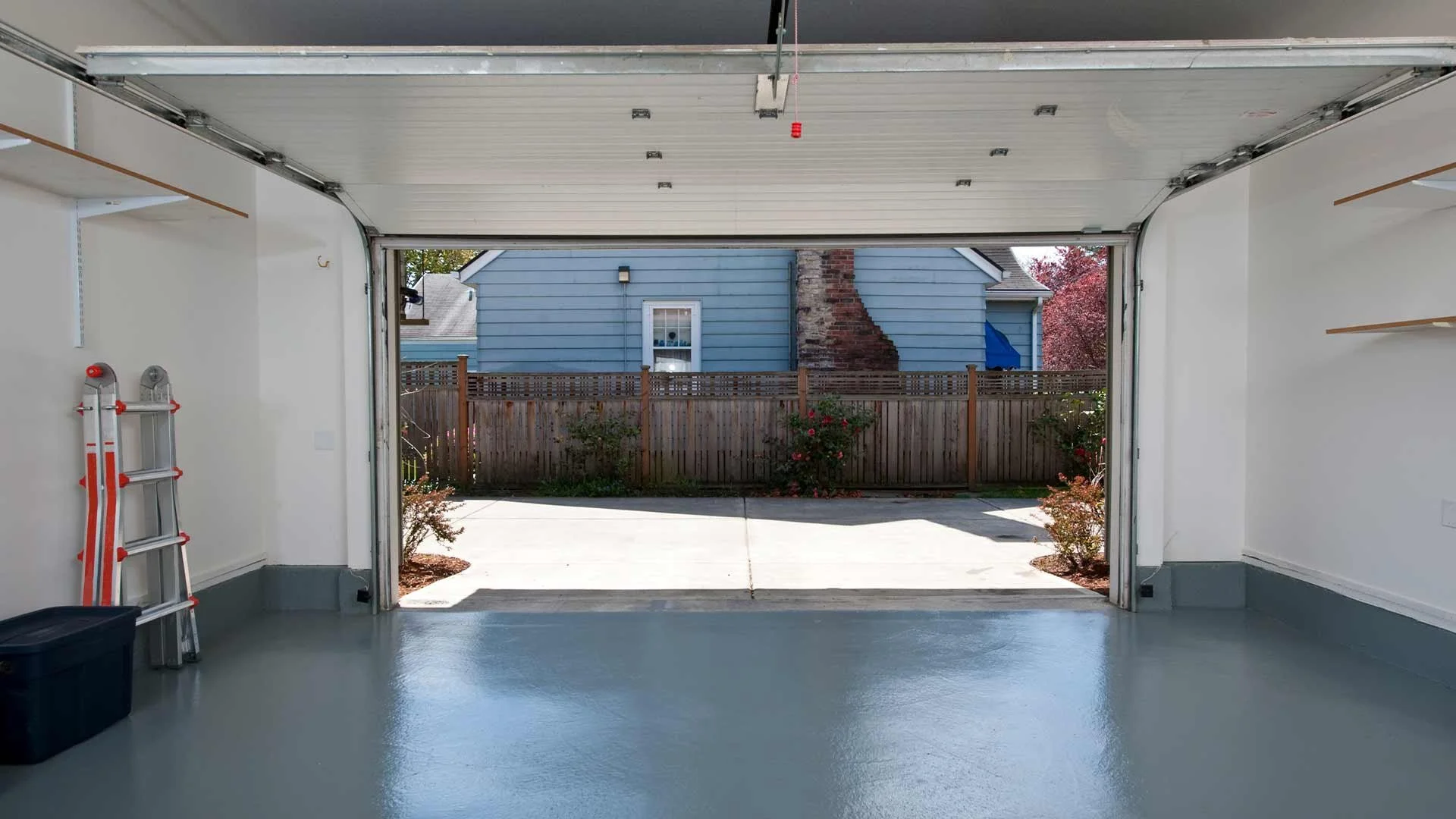 An open garage door showing a clean, organized garage interior, with a ladder and storage shelves on the left side, and a view of a fenced backyard with a blue house and plants.