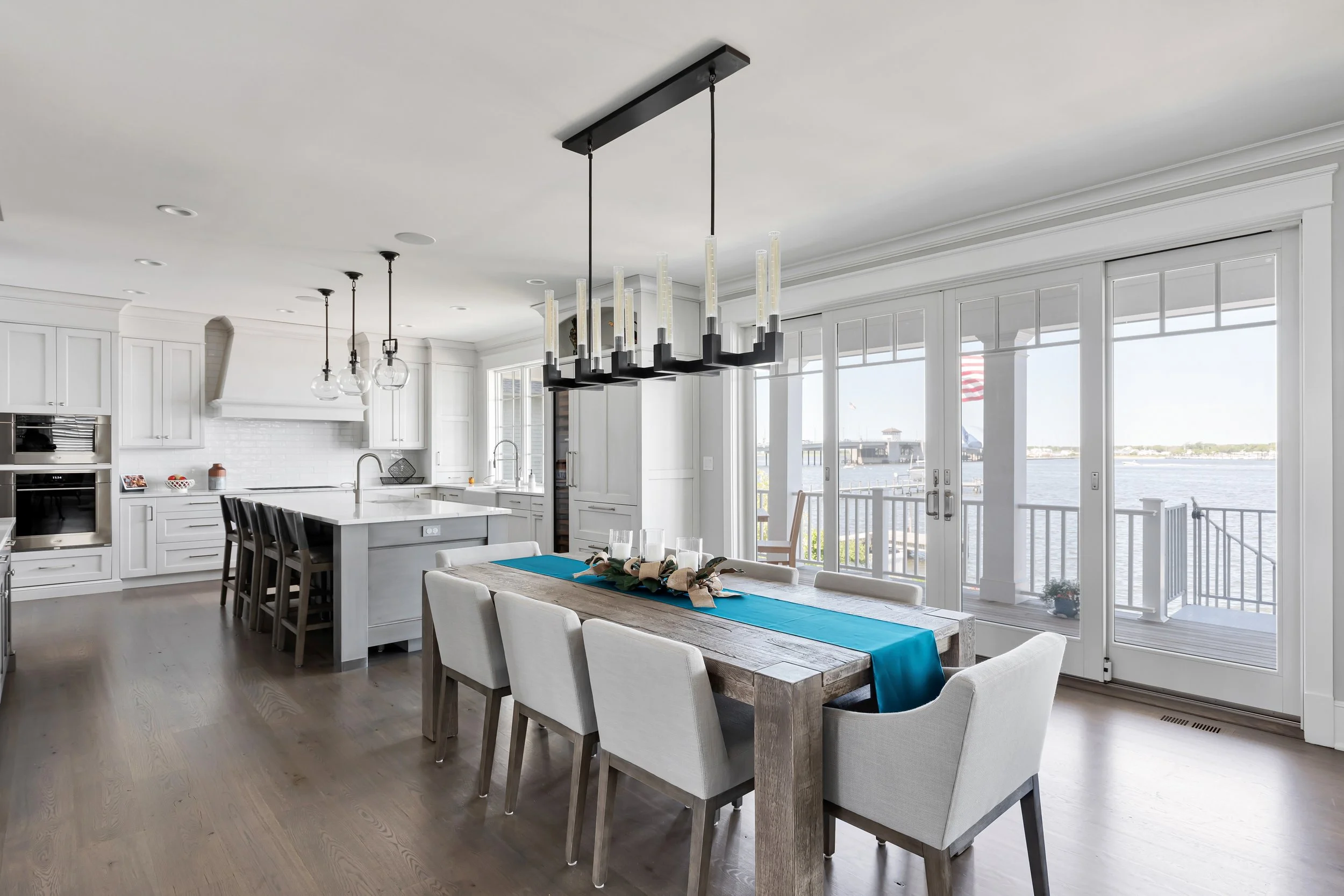 Bright, modern kitchen and dining area with white cabinetry, a large island with stools, and a wooden dining table with chairs. Floor-to-ceiling windows and sliding glass doors offer a view of a waterfront scene with a dock and American flag outside.