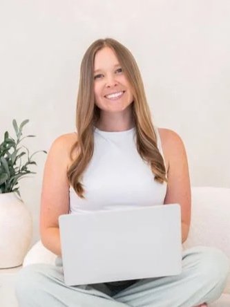 A woman with long blonde hair smiling, wearing a white blazer, white top, blue jeans, and a brown belt, standing in a bright white room next to a desk with a mug and books.