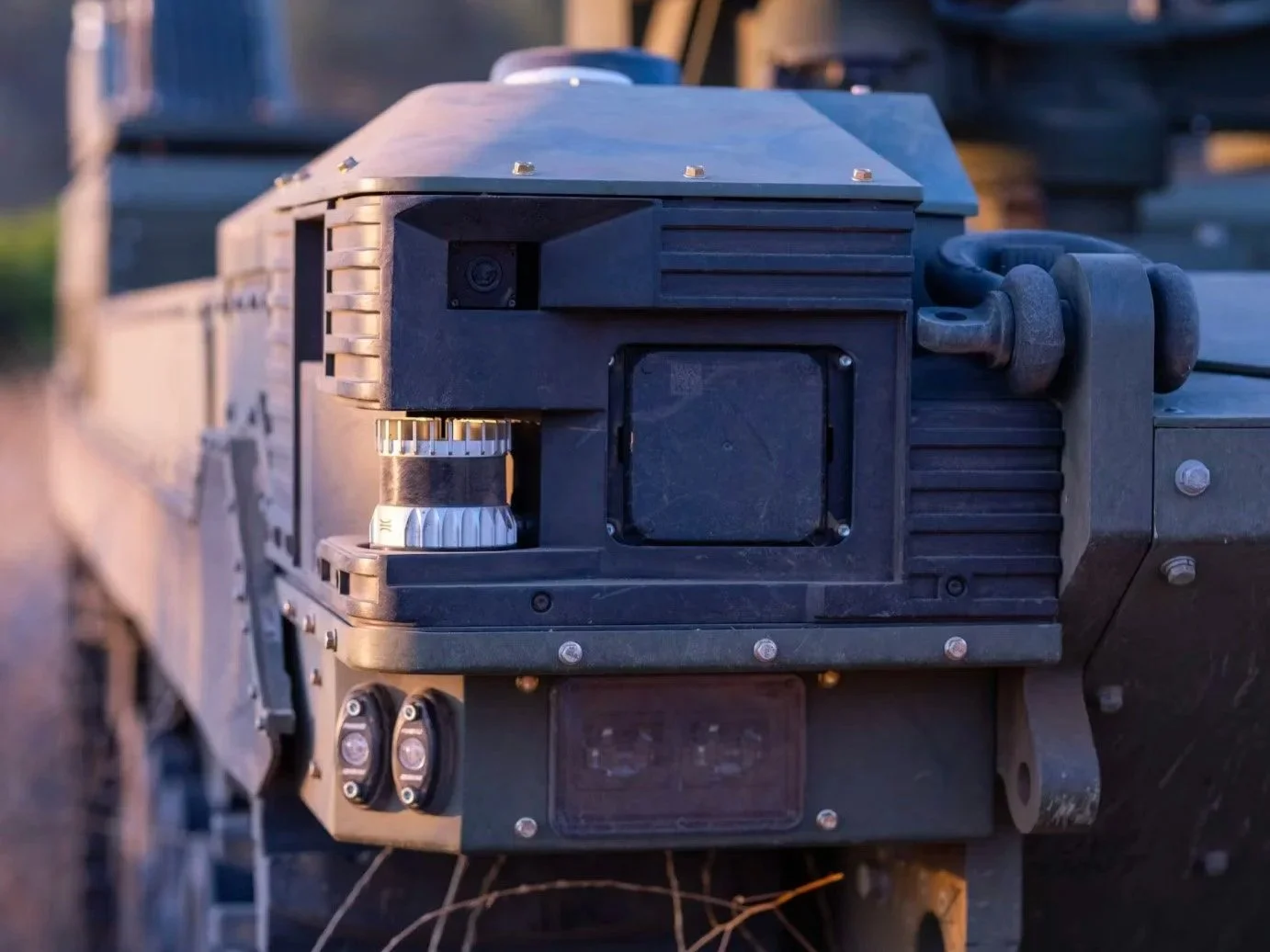 Close-up of a black military or tactical equipment device with a shiny metallic component, installed on a vehicle or machinery, during daytime.