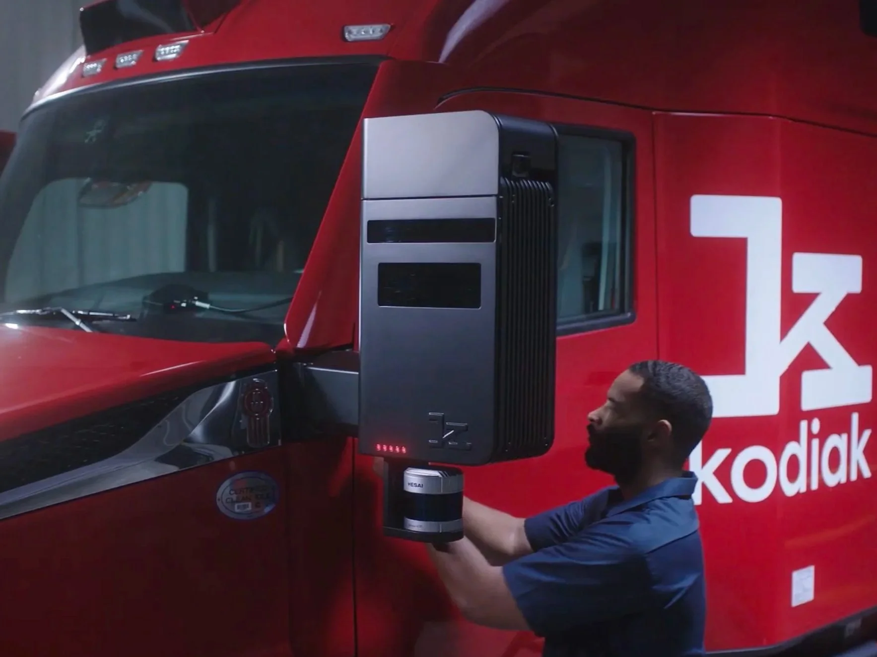 A man in a blue shirt demonstrates a large industrial machine in front of a red vehicle with the 'Kodiak' logo, likely at a trade show or exhibition.