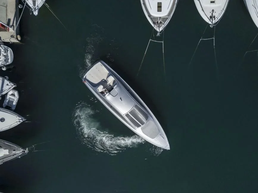 Aerial view of a sleek silver motorboat moving through a marina, surrounded by anchored sailboats.
