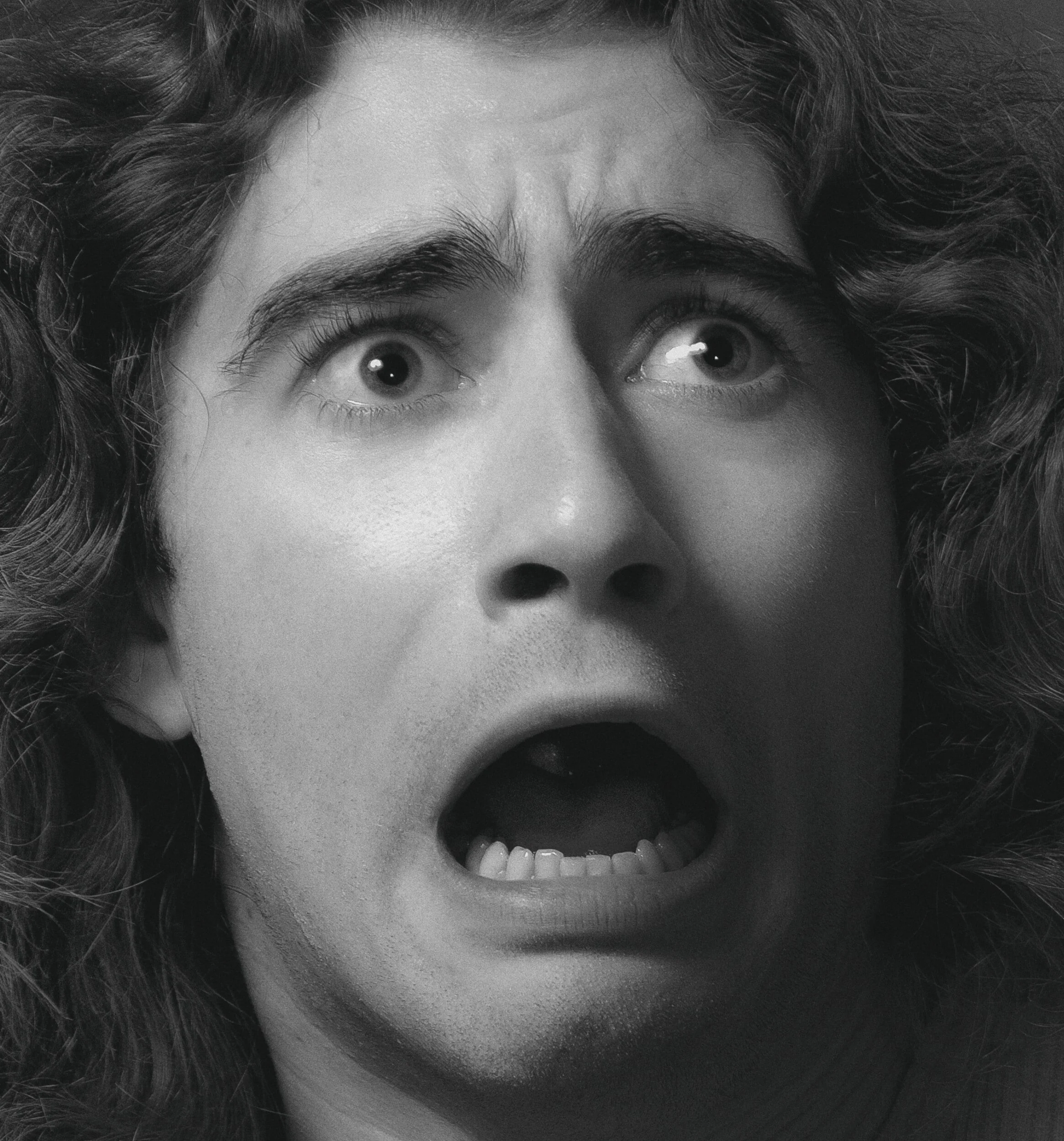 A black and white close-up of a person with curly hair expressing surprise or shock, mouth open and eyes wide