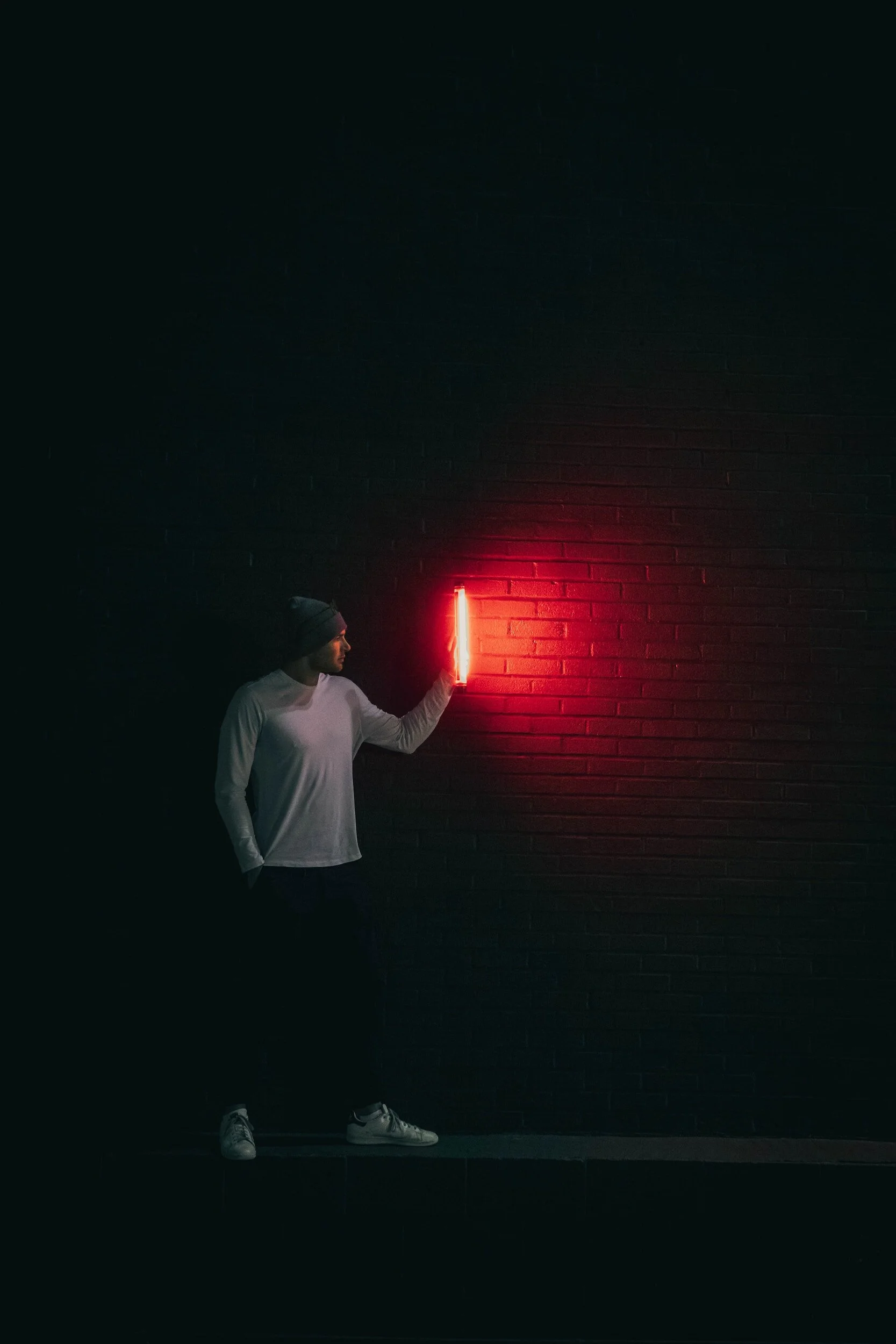 Person wearing a beanie and white long sleeve shirt, standing in front of a dark brick wall, touching a vertical red neon light with their hand.