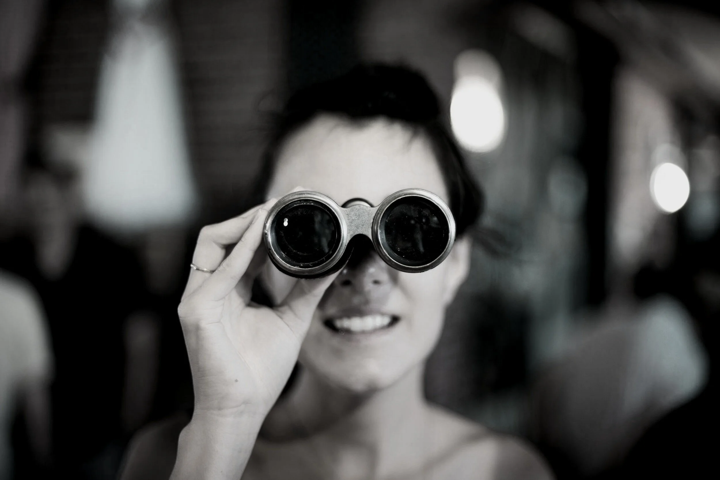 A woman holding binoculars up to her eyes and smiling, in black and white photo