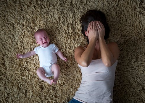 A crying baby lying on a yellow shaggy carpet next to a woman who is lying on her back with her hands covering her face.
