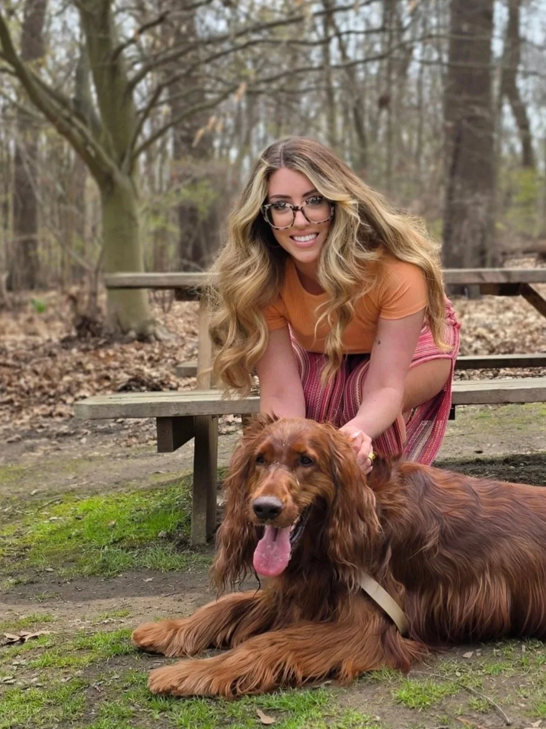 A young woman with long, wavy blonde hair and glasses sits on a picnic bench beside a large, long-haired red Irish setter dog with floppy ears and a pink tongue. They are outdoors in a wooded area with trees.