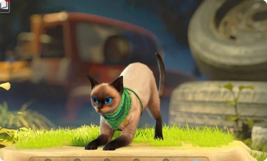 A Siamese cat with bright blue eyes, wearing a green scarf, crouching on a patch of green grass outdoors with a blurred background featuring a red vehicle, a tire, and some stone structures.