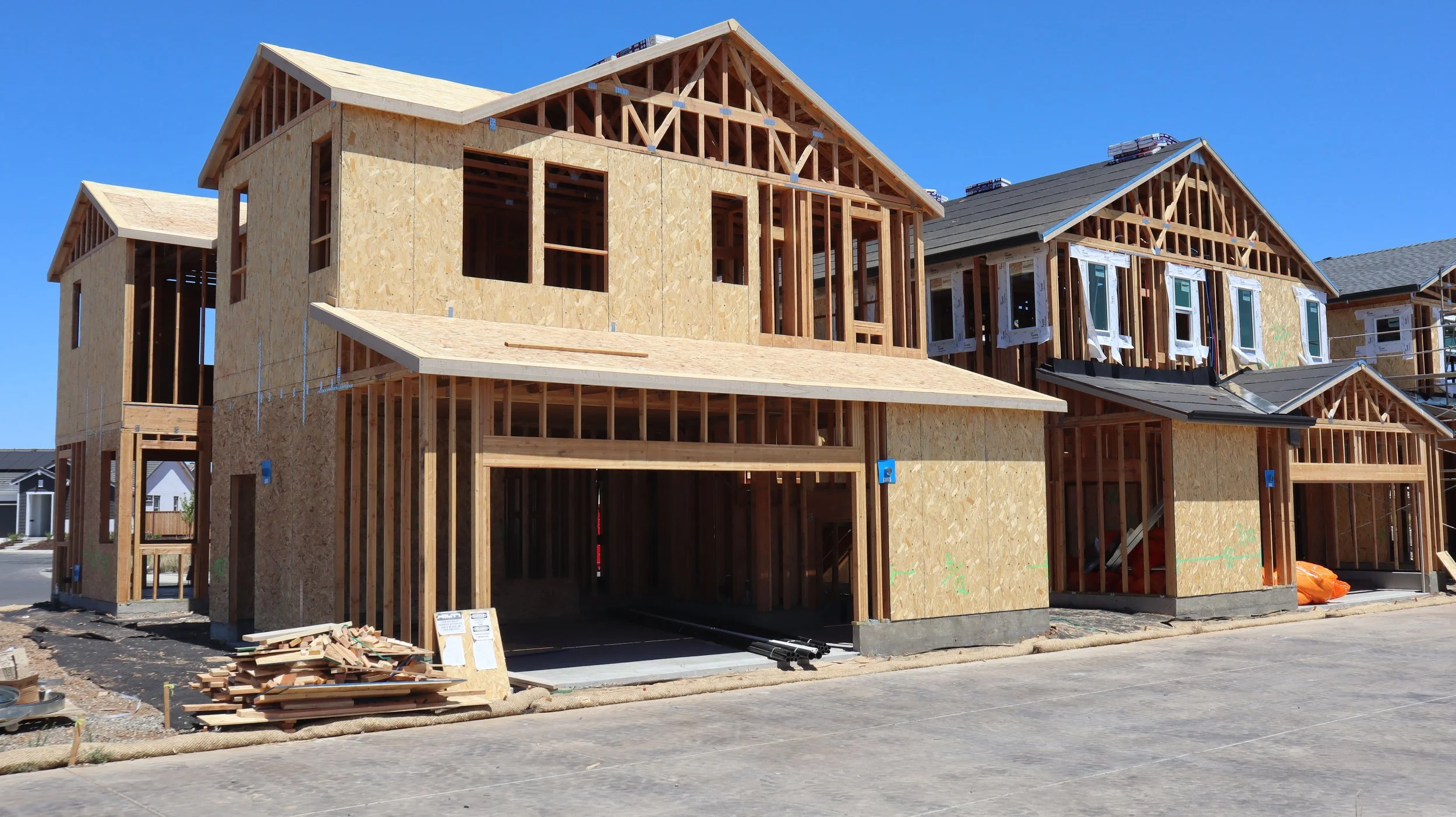 A house under construction with wooden framing and plywood sheathing, multiple gables, and a clear blue sky.