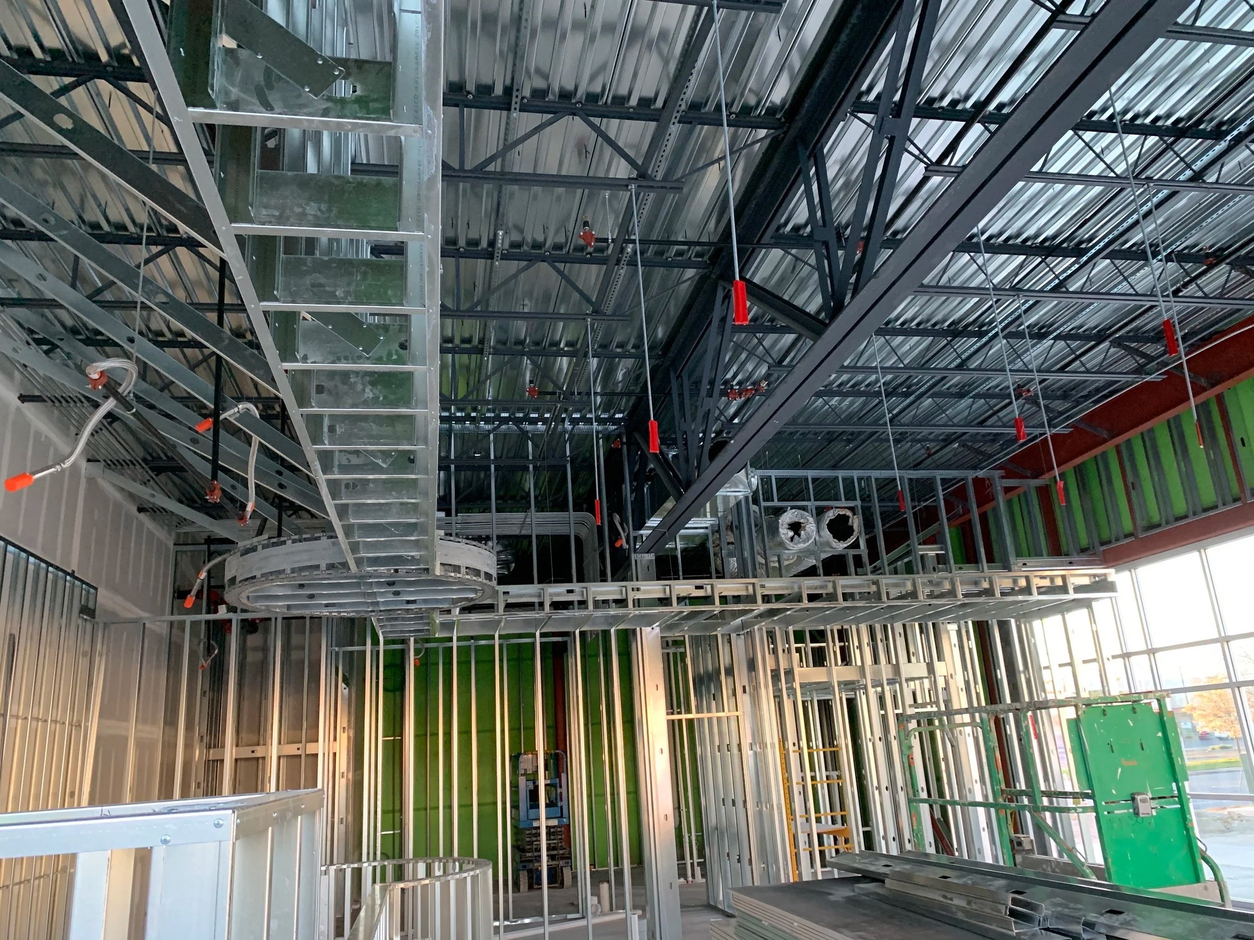 Interior view of a building under construction, showing metal framing and piping, with some green wall panels, a ladder, and construction equipment.