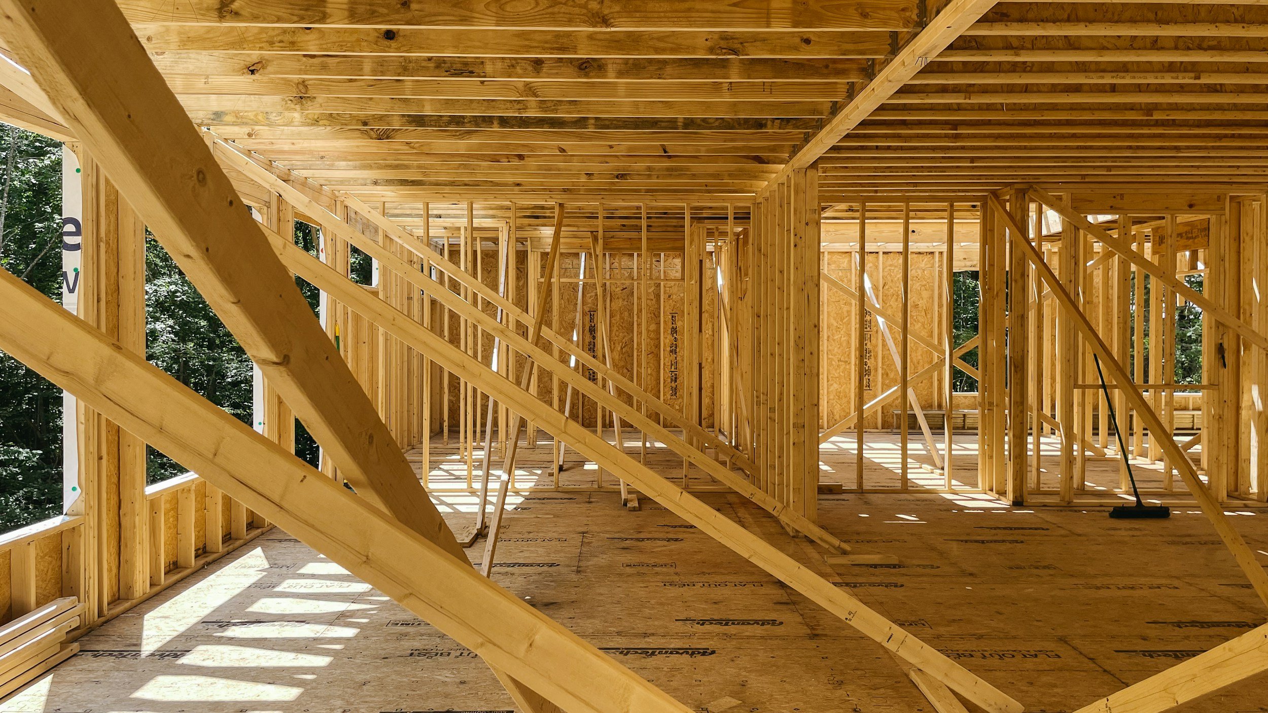 Interior view of a wooden residential building under construction, showing framing with vertical and horizontal beams, and an unfinished ceiling with exposed roof rafters.