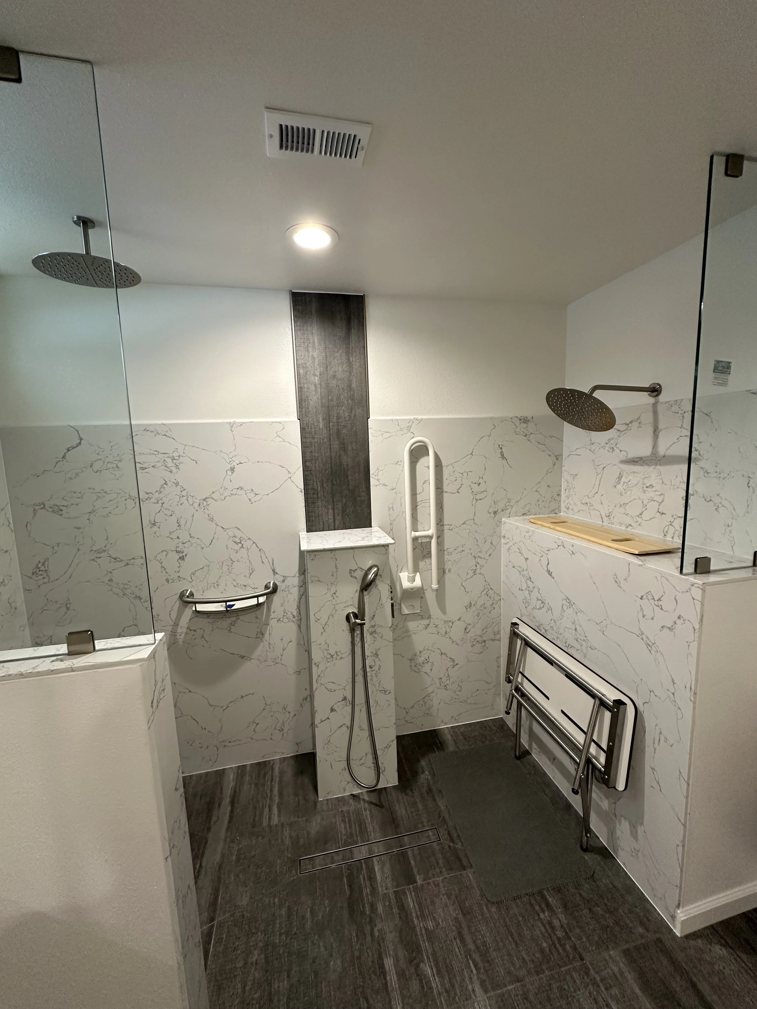 Accessible shower with two handheld showerheads, marble-style wall tiles, a small bench, grab bars, a wooden amenity tray, a mirror, and dark wood-look flooring.