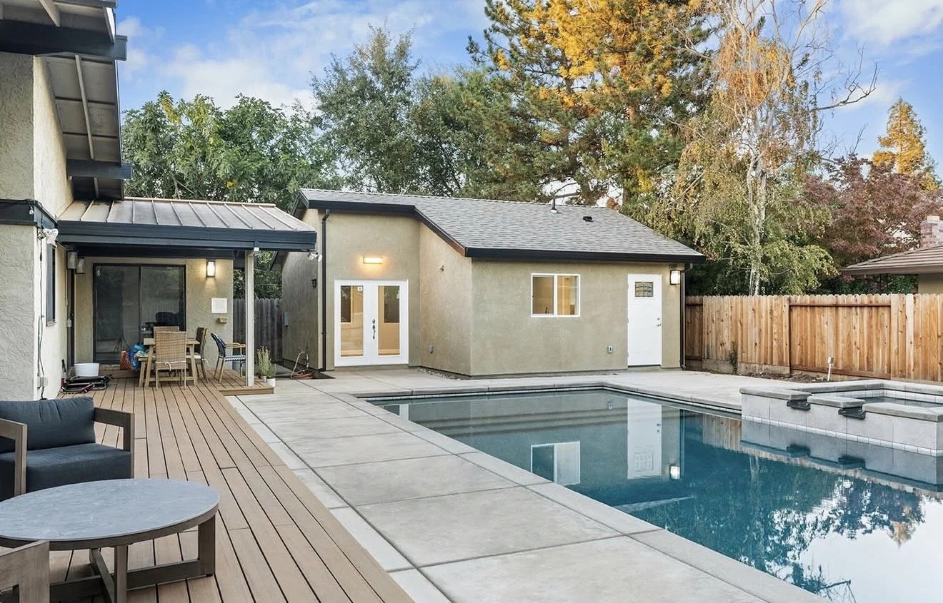 Residential backyard with a swimming pool, wooden deck area with outdoor furniture, and a small beige building with doors and windows, surrounded by trees and a wooden fence.