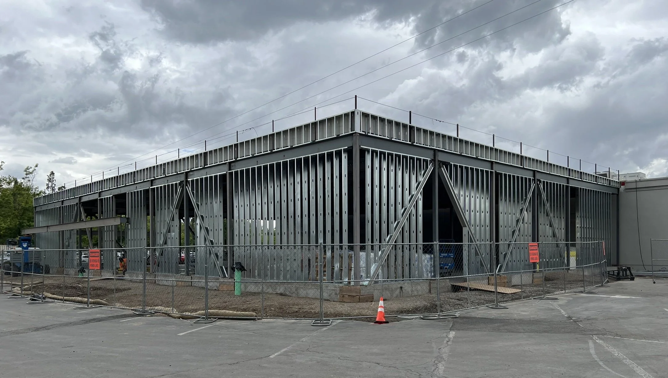 A building under construction with metal framing, surrounded by a chain-link fence and orange construction signs, under a cloudy sky