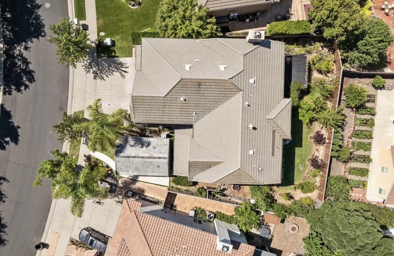 An aerial view of a backyard, showing a house with a beige roof, surrounded by a garden with trees and shrubs, a wooden deck, and neighboring houses with tiled roofs.