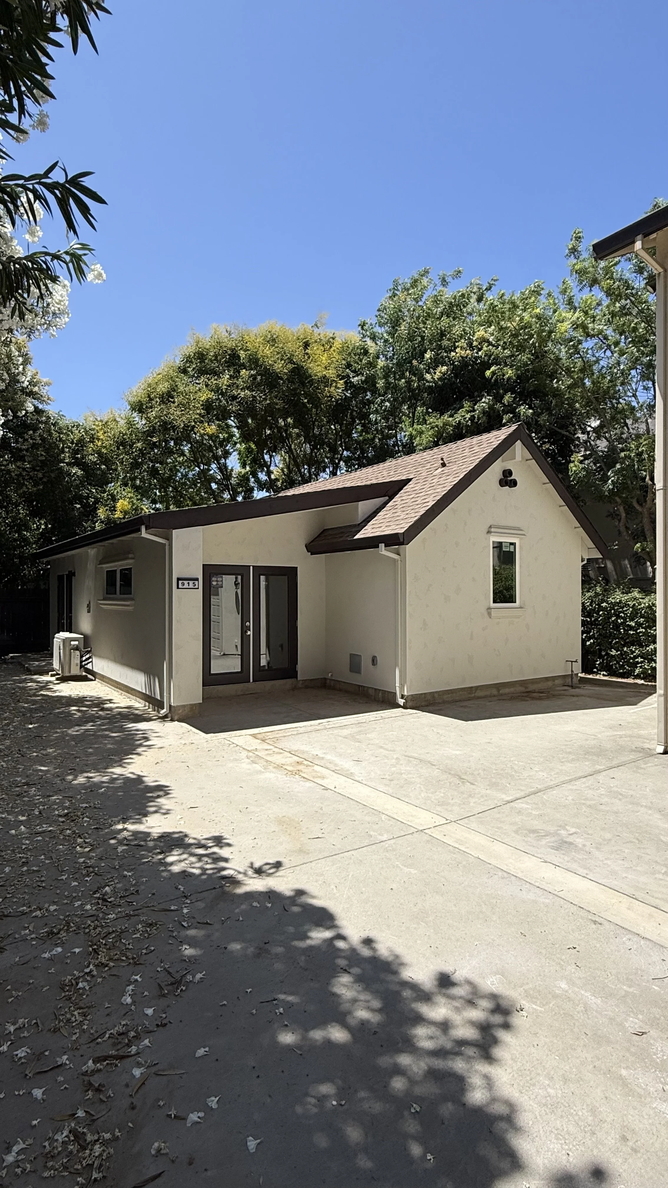 Small white house with a sloped roof, glass door, and a small window, surrounded by trees and a concrete driveway under a clear blue sky.