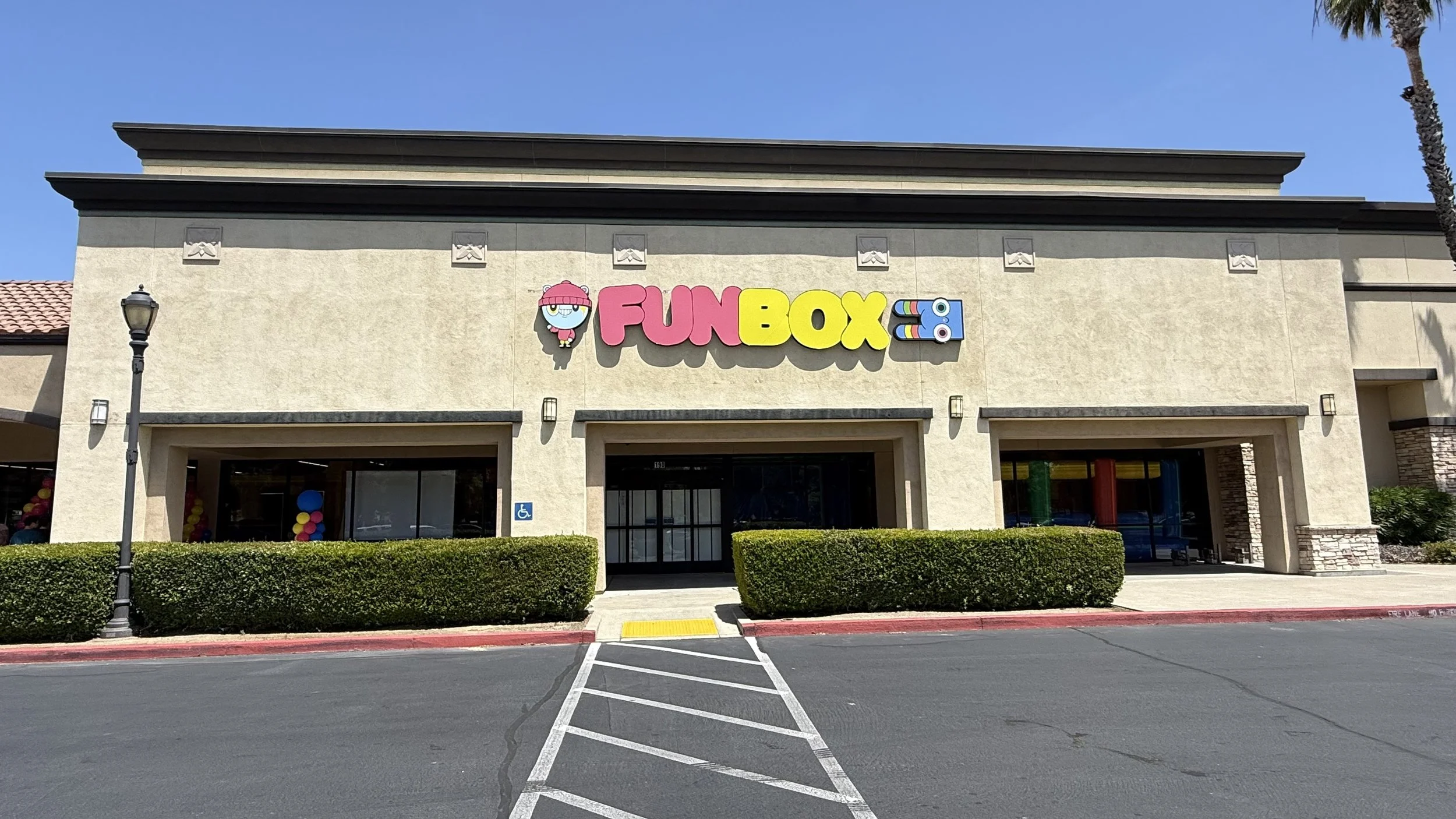 Building with a sign that says 'Fun Box' next to a cartoon character, palm trees, and bushes in the parking lot.