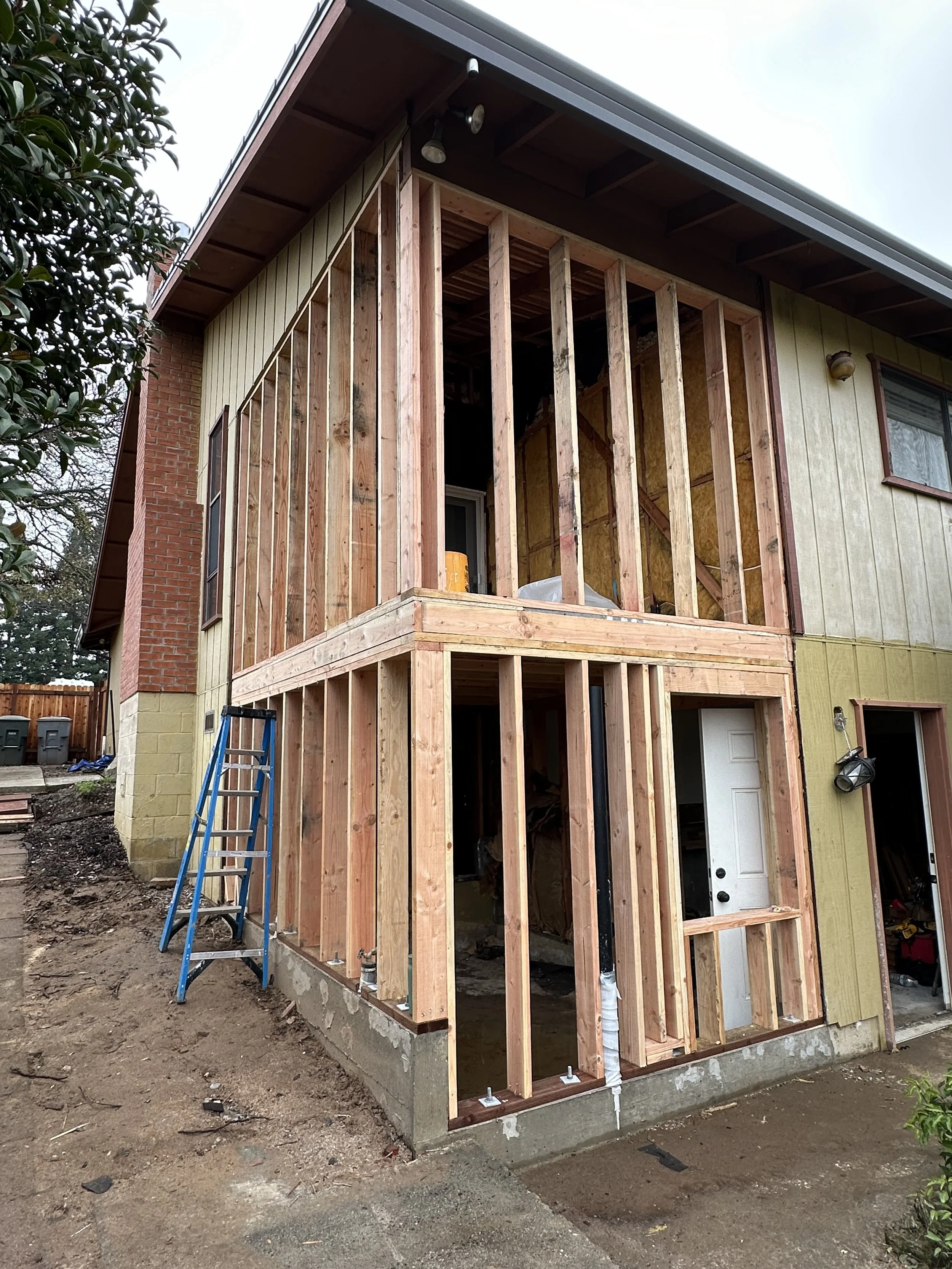 The image shows a house under construction, with a wooden frame added on the front side. The house has yellow siding on one part, a brick chimney, and a gray roof. There is a ladder leaning against the house, and construction materials are visible inside the frame.