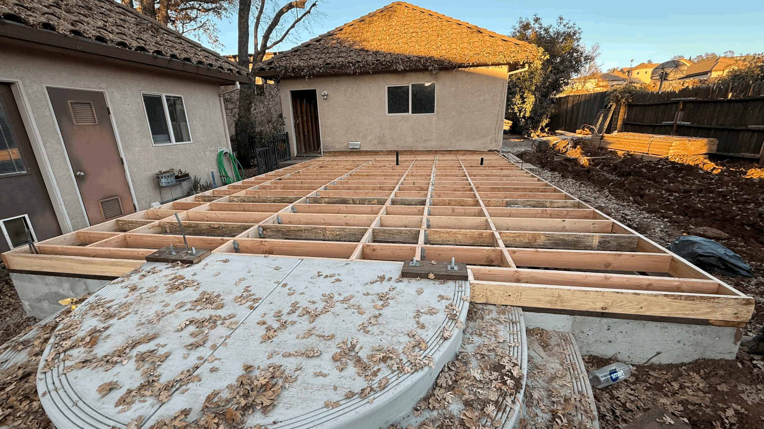 Photo of a house under construction with a wooden framework for a new deck or patio area in the backyard. The framework is made of wooden beams and there are two neighboring houses visible, one with a tile roof. The yard has some soil and rocks, and the sun is setting, casting a warm glow.