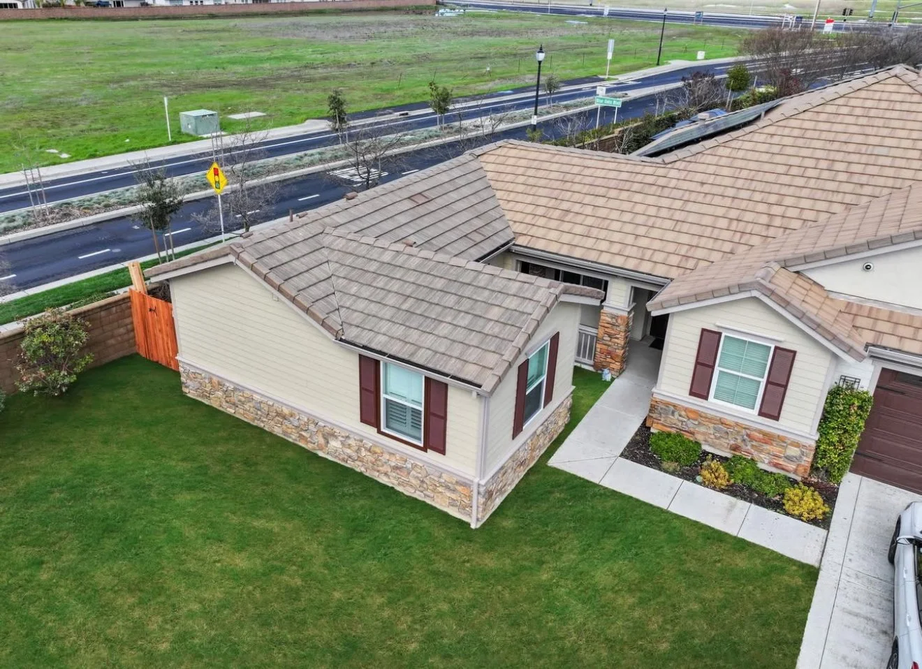 Aerial view of a beige house with stone accents, a brown tiled roof, and a small front yard with landscaped bushes. Next to it is a driveway and a neighboring house with similar features. A street with trees and streetlights runs behind the houses.