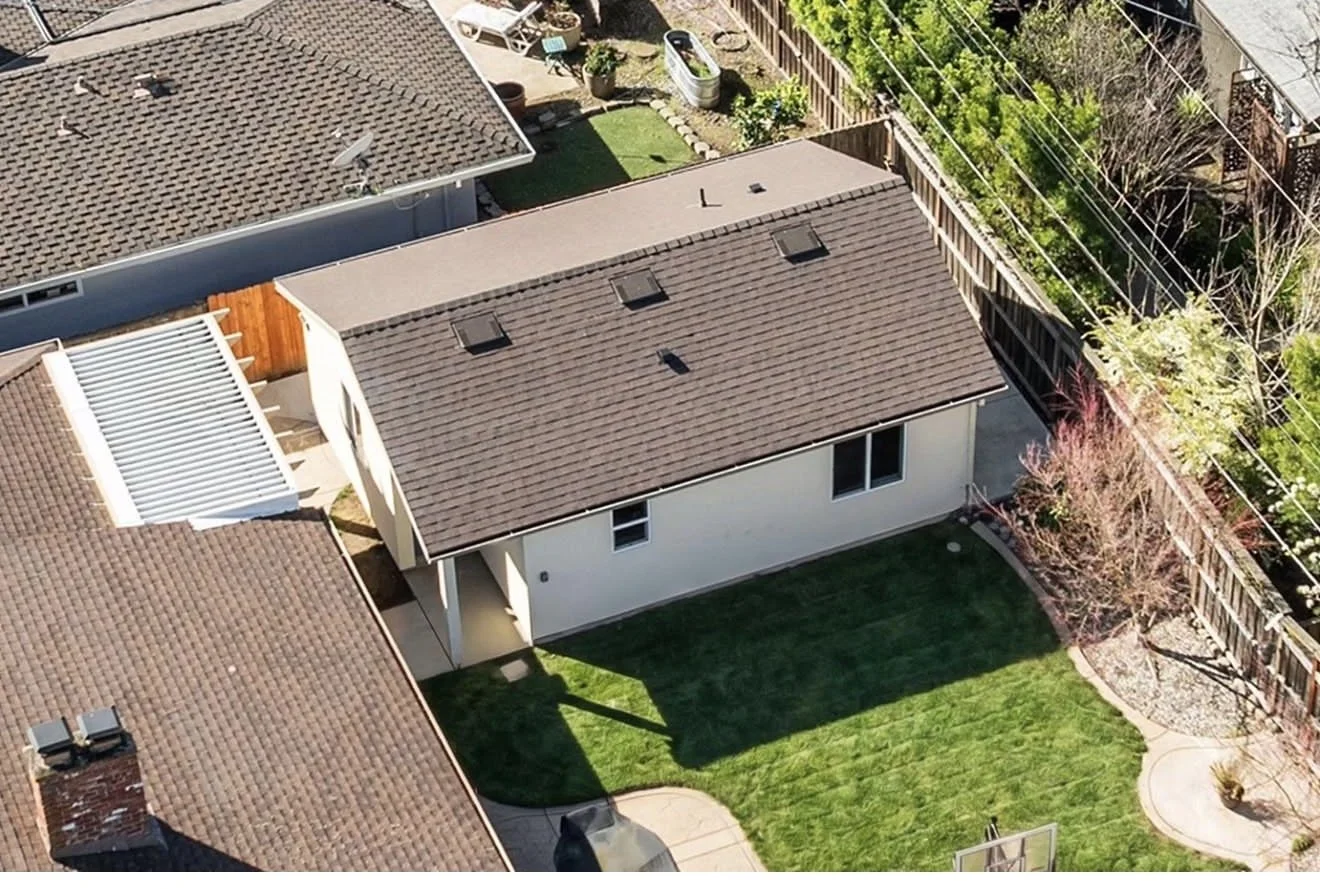 Aerial view of a backyard with a house, green lawn, patio area, and surrounding neighboring rooftops and fences.