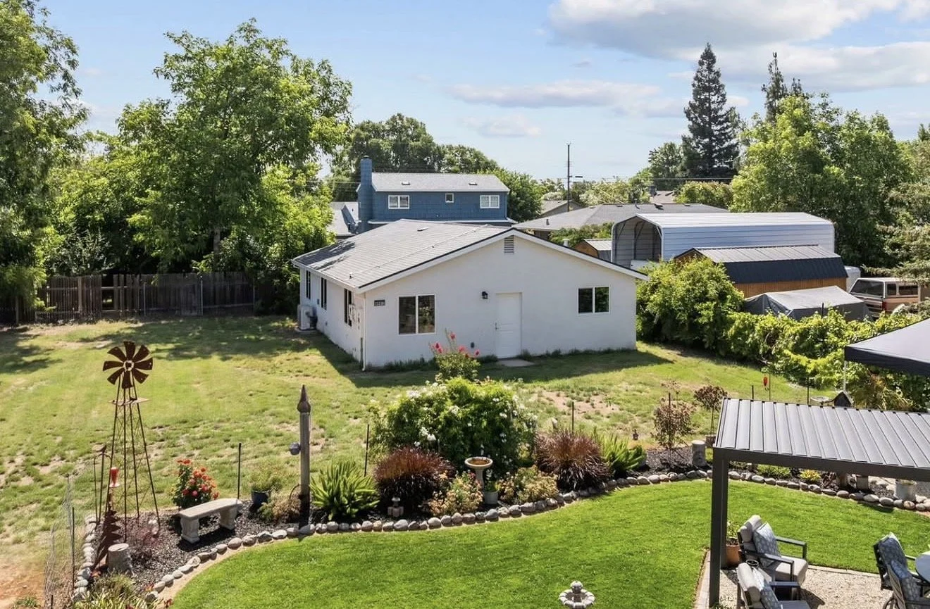 Backyard view with a white house, trees, garden flowers, a windmill decoration, and patio furniture under a gray roofed pergola.