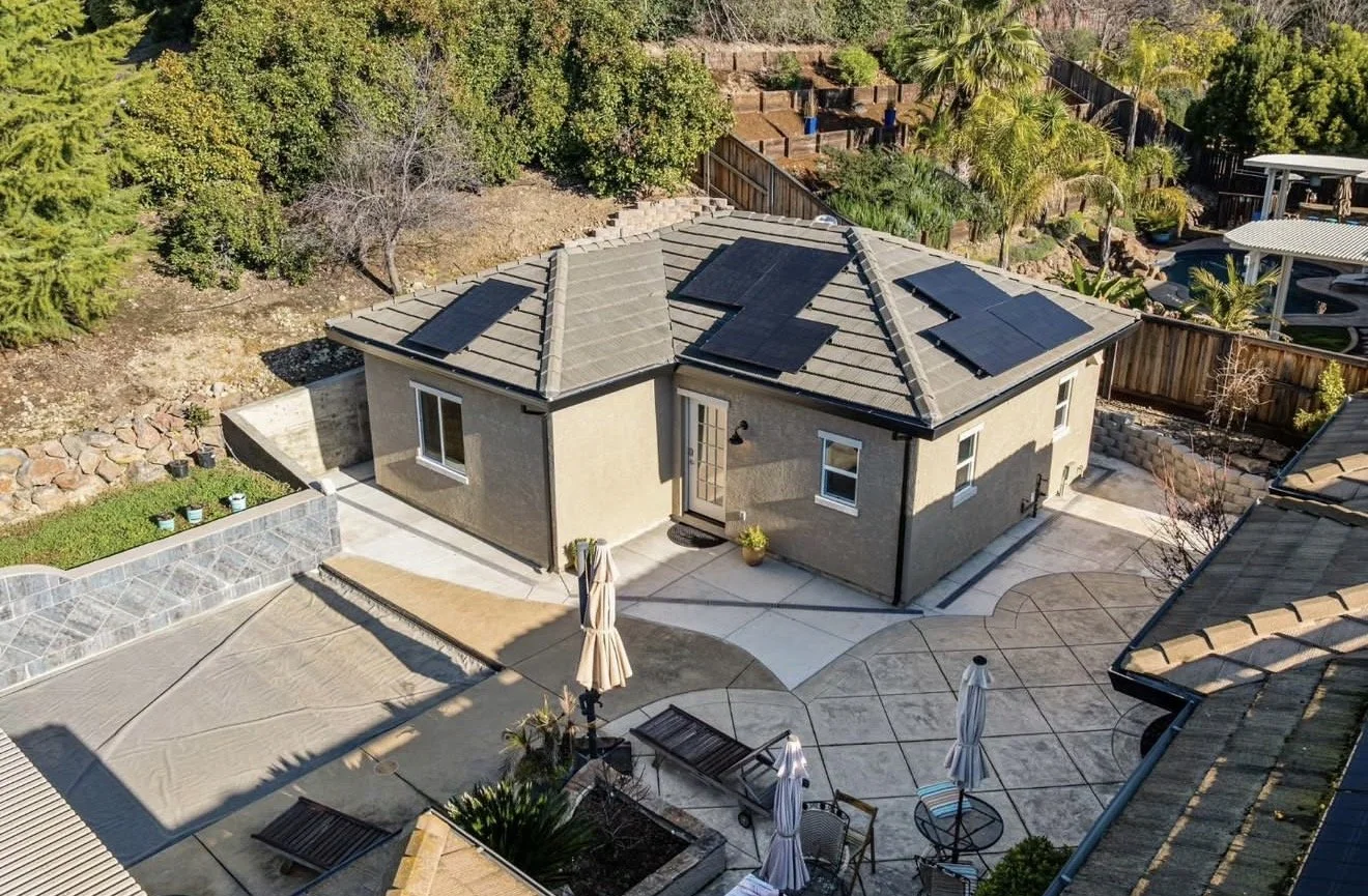 A backyard with a beige house equipped with solar panels on the roof, a paved patio area with outdoor furniture, an umbrella, and a lawn with a small garden bed surrounded by stone wall.