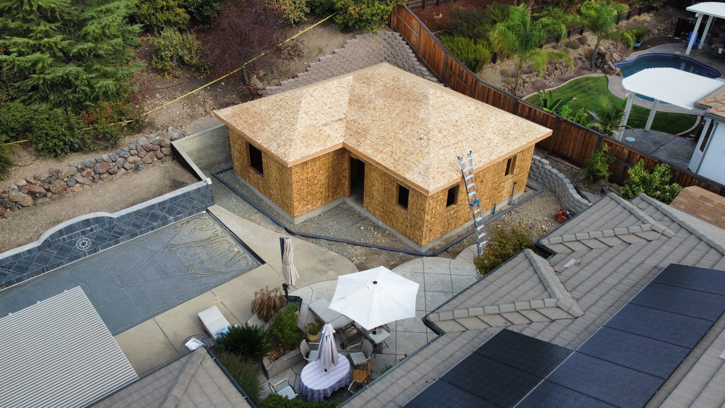 A residential backyard with a house under construction, showing a plywood wall and roof framing. The backyard includes a pool with a cover, outdoor furniture with umbrellas, a tiled patio, and landscaped areas with trees and shrubs.