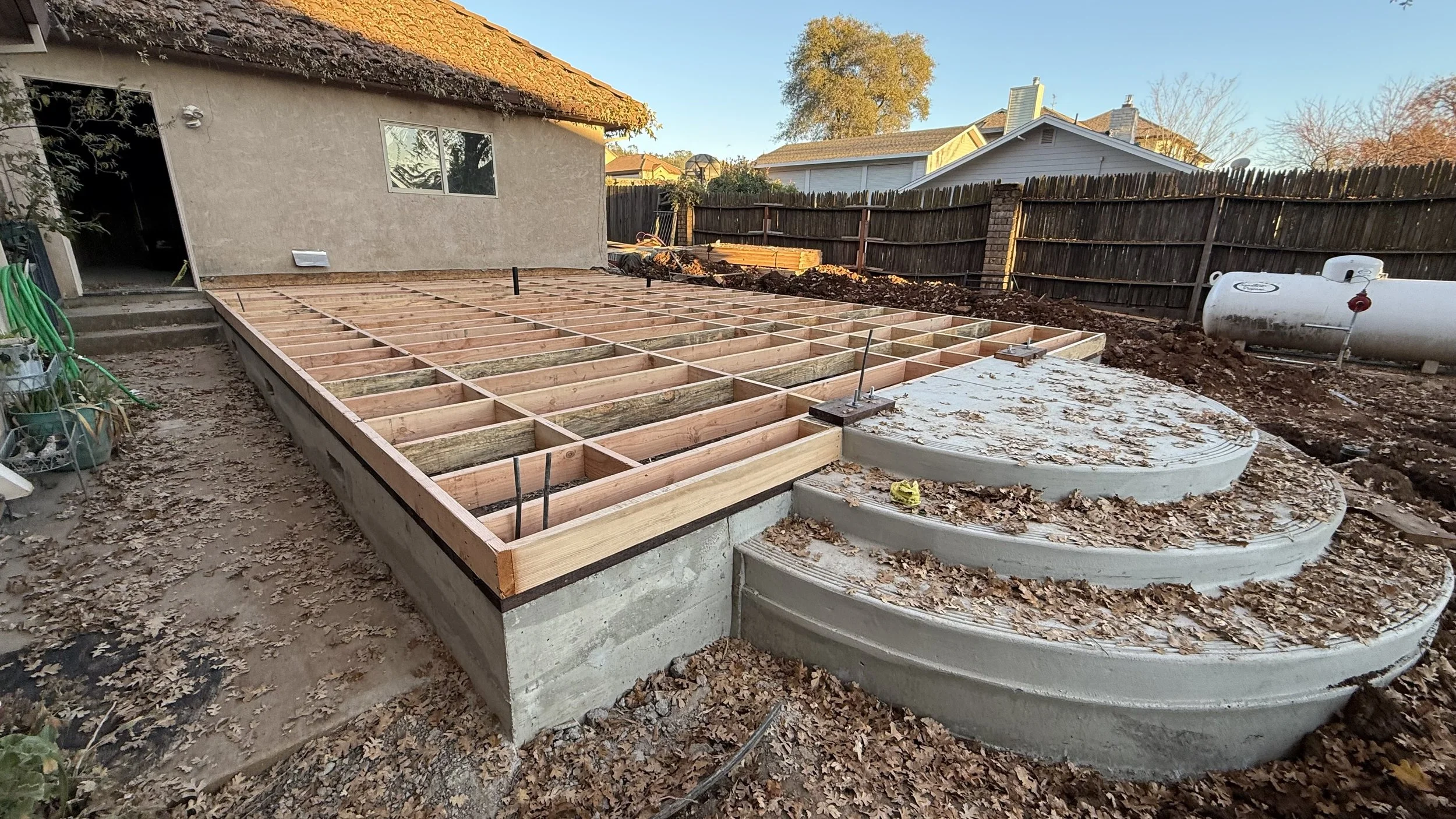 Backyard construction site with a wooden framework for a deck or patio, concrete stairs, large gas tank, and surrounding fallen leaves.