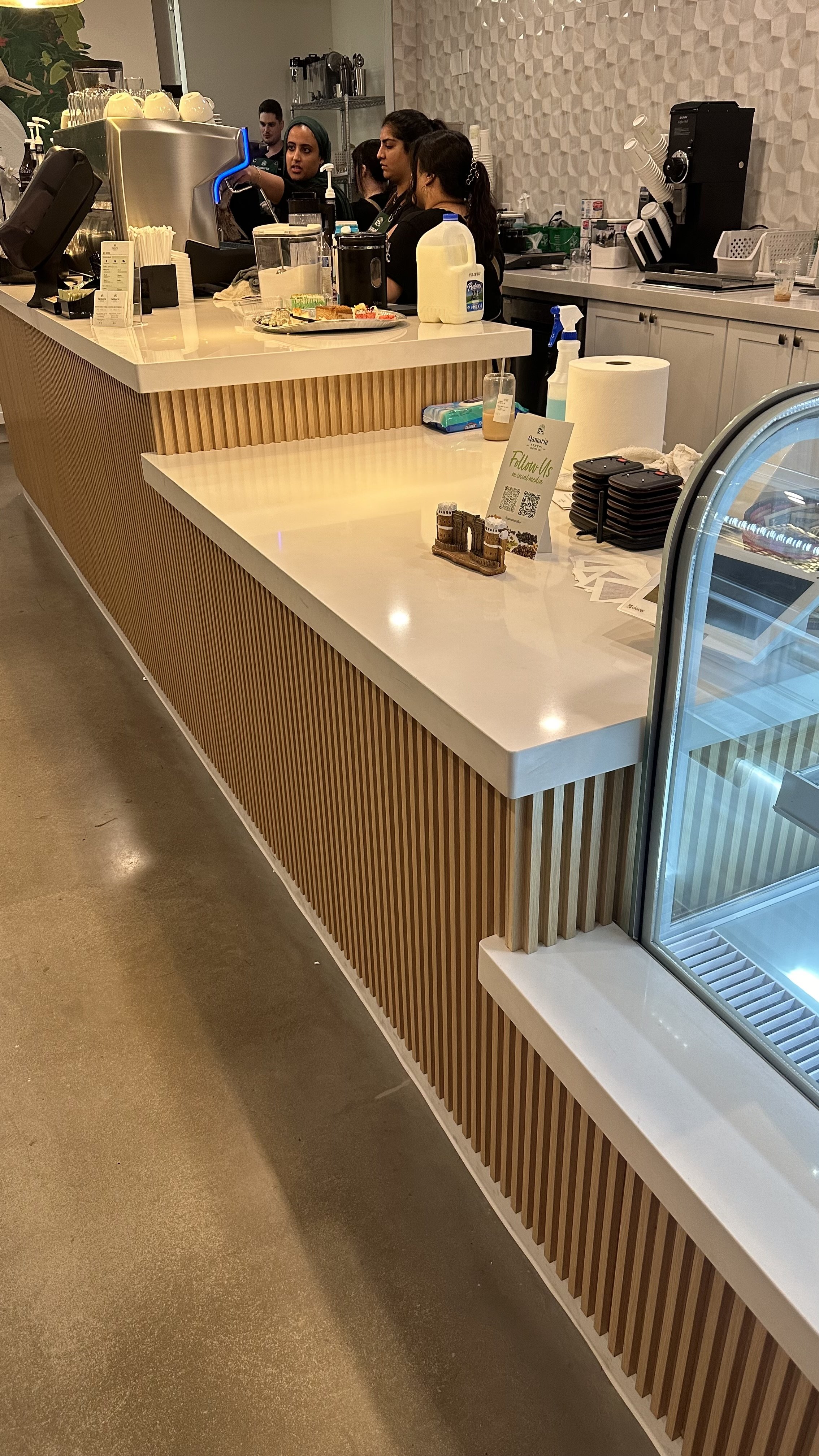 Interior of a modern cafe or coffee shop with a wooden slatted counter and a glass display case. Several employees work behind the counter, with one pouring a drink from an espresso machine. Items like milk, paper towels, and trays are on the countertop.