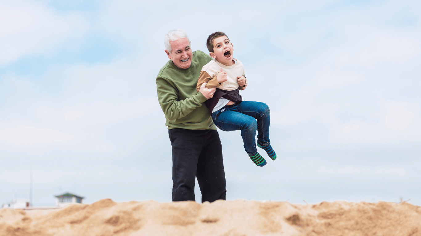 a grandfather and grandson who are part of a multigenerational family are playing on a sand dune, laughing