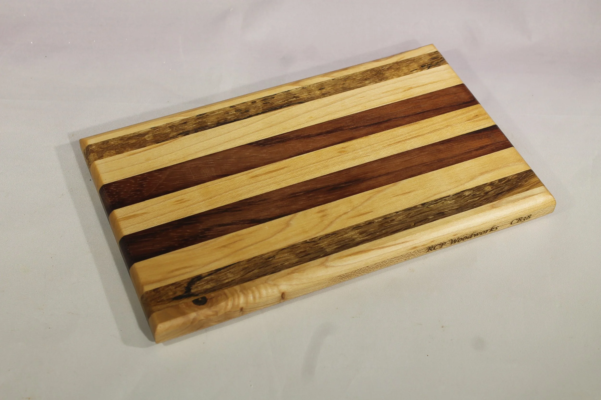 CB38  -   Cheese & Cutting board.     $60

5 5/8” wide by 9 1/8” long by 5/8” thick.
Nine segments of wood – Maple, Oak, Maple, Bubinga, Maple, Buginga, Maple, Oak, Maple.

Finished with cutting board oil.
