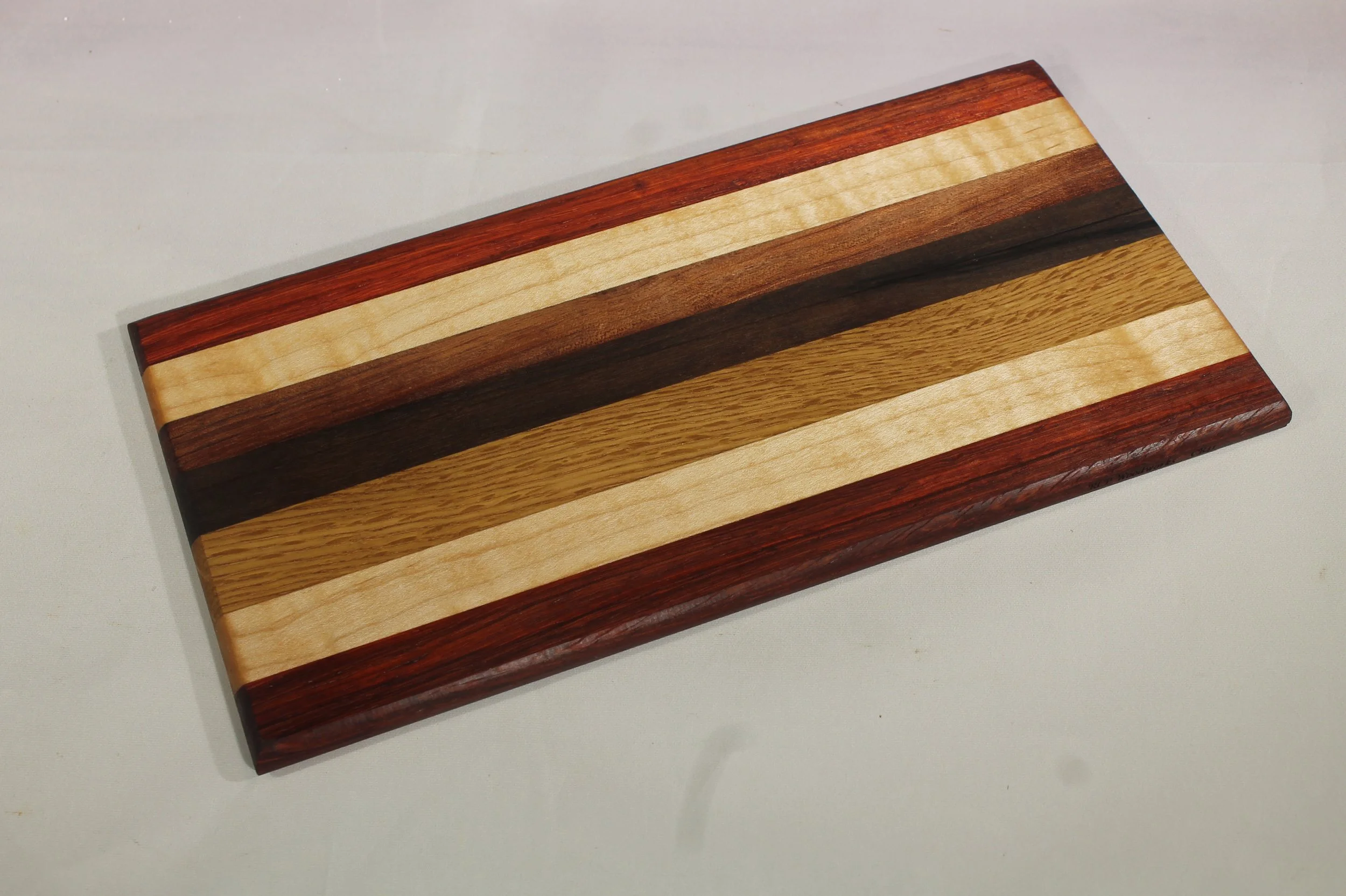 CB41A - Cheese & Cutting board.

5 3/8” wide by 10 ½” long by ½” thick.
Seven segments of wood – Bubinga, Maple, Oak, Walnut, Bubinga, Maple, Sapele.

Finished with cutting board oil.      $55