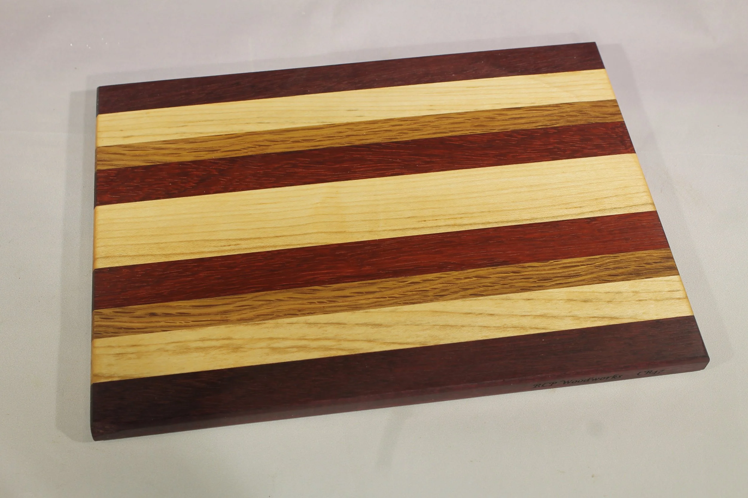 CB47 -  Cheese & Cutting board.

6 ¾” wide by 9 3/8” long by 11/16” thick.
Nine segments of wood – Purpleheart, Maple, Oak, Paduk, Maple, Paduk, Oak, Maple, Purpleheart.

Finished with cutting board oil.       $65
