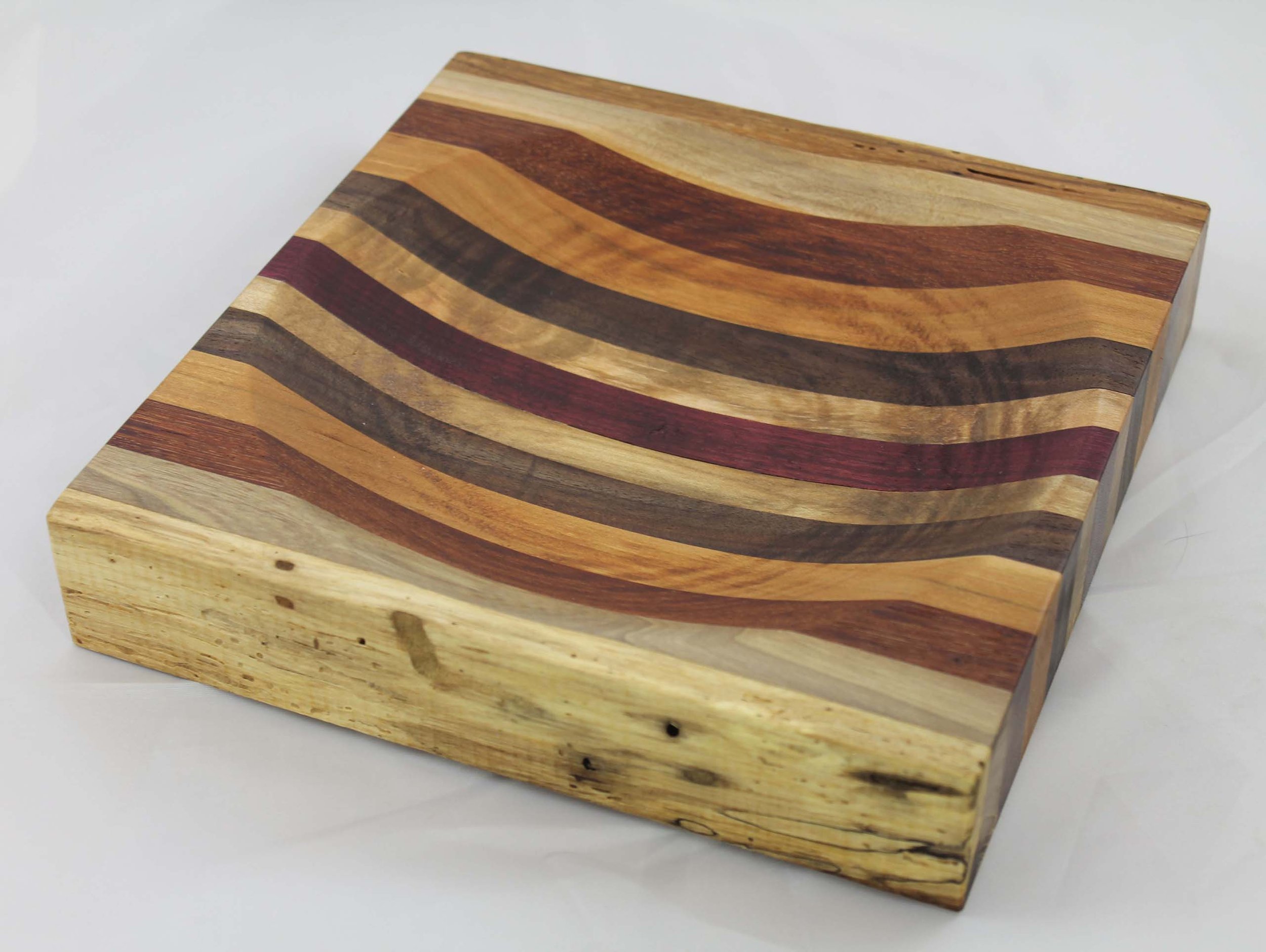 CB 29 - Ulu knife cutting board.  This cutting board measures 8 ⁷⁄₈”W x 8 ⁷⁄₈”L x 1⁵⁄₈” H.   It has13 segments – Walnut / Elm / Jatoba / Cherry / Walnut / 
Poplar / Purpleheart / Poplar / Walnut / Cherry / Jatoba / Elm / Walnut. 
$45