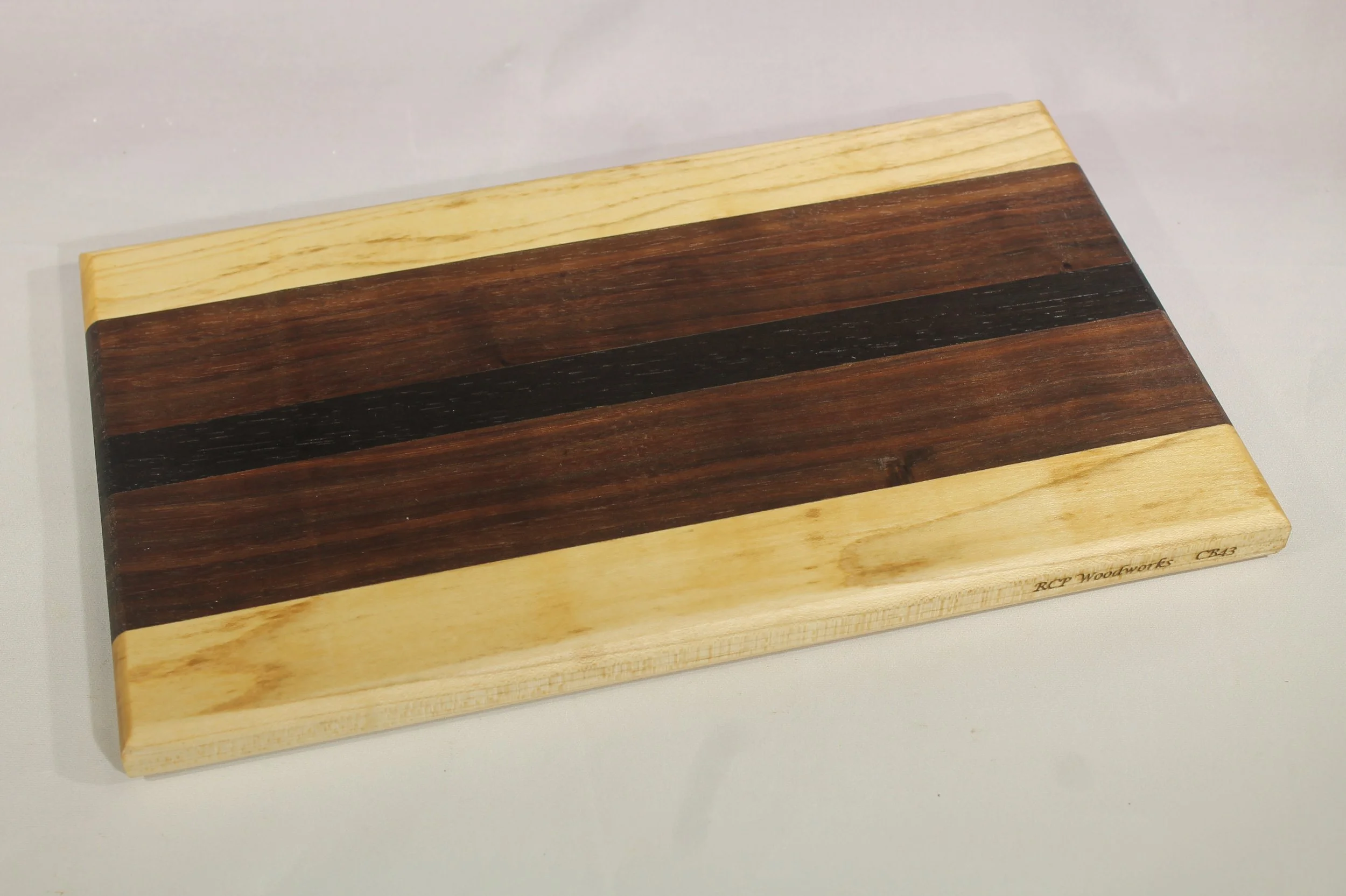 CB43  -  Cheese & Cutting board         $55

6 ½” wide by 11” long by ¾” thick.
Five segments of wood – Maple, Walnut, Wenge, Walnut, Maple

Finished with cutting board oil.
