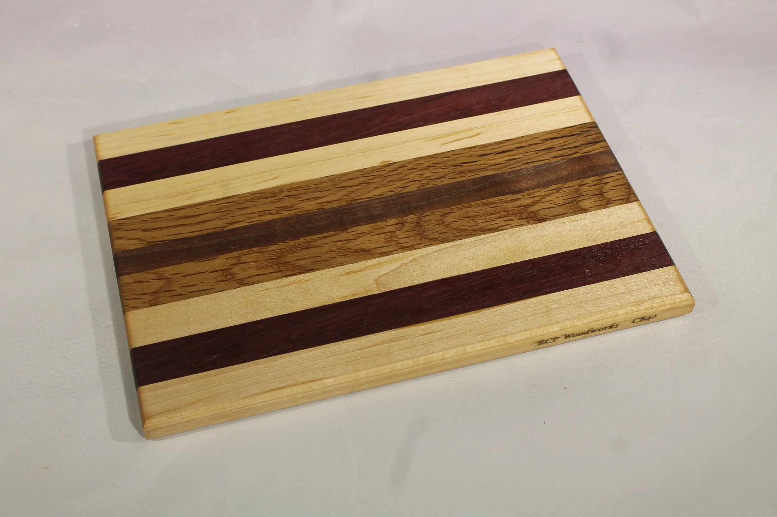 CB 42A - Cheese & Cutting board.

6 3/8” wide x 9” long by 11/16” thick.
Nine segments of wood – Maple, Purpleheart, Maple, Oak, Walnut, Oak, Maple, Purpleheart, Maple.

Finished with cutting board oil.        $65
