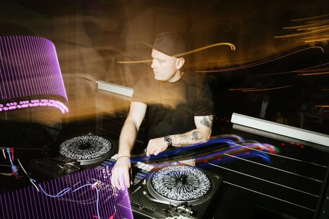 Ein DJ in Schwarz beim Auflegen, mit Turntables und Mischpult, in einem dunklen Club mit Lichteffekten.